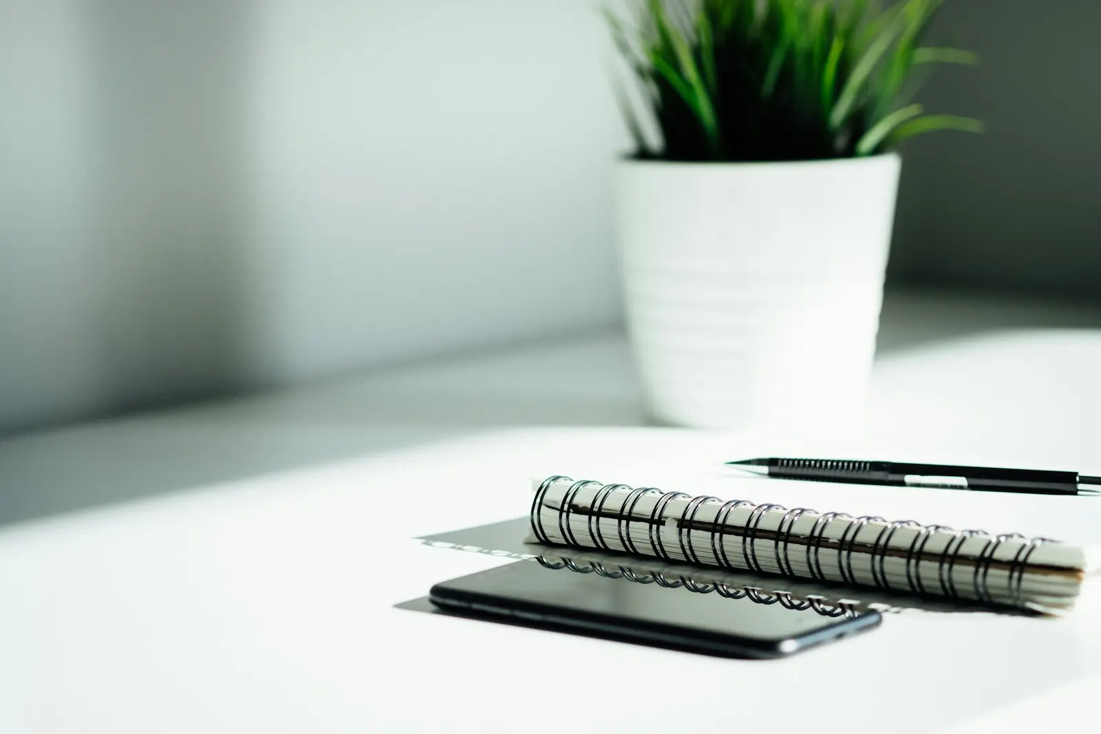 A smartphone lying on a white desk beside a spiral notebook and a pen, with a potted plant in the background