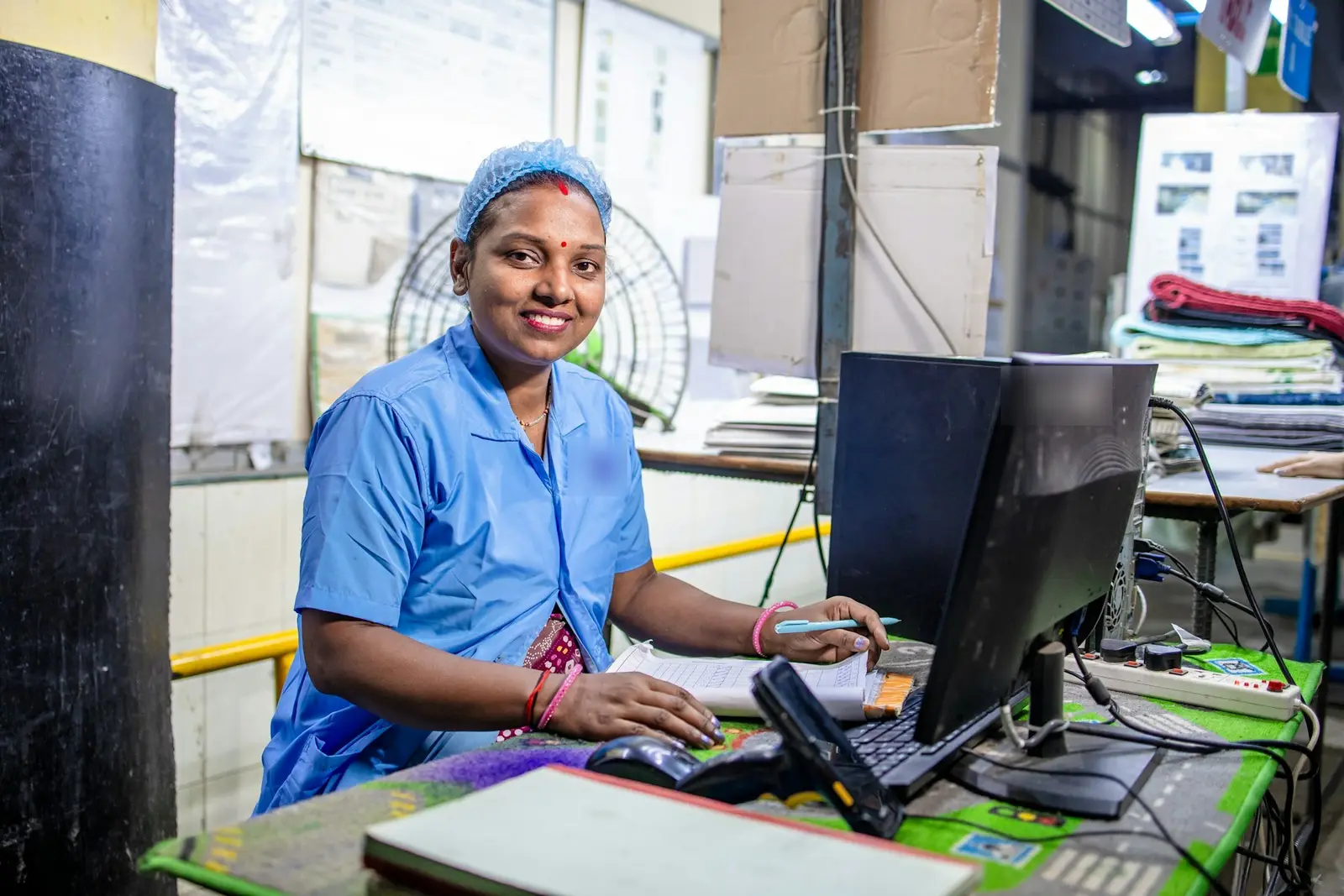 A smiling woman in a blue work uniform and hair net sitting at a computer desk with a pen and a printed sheet, in a busy workroom