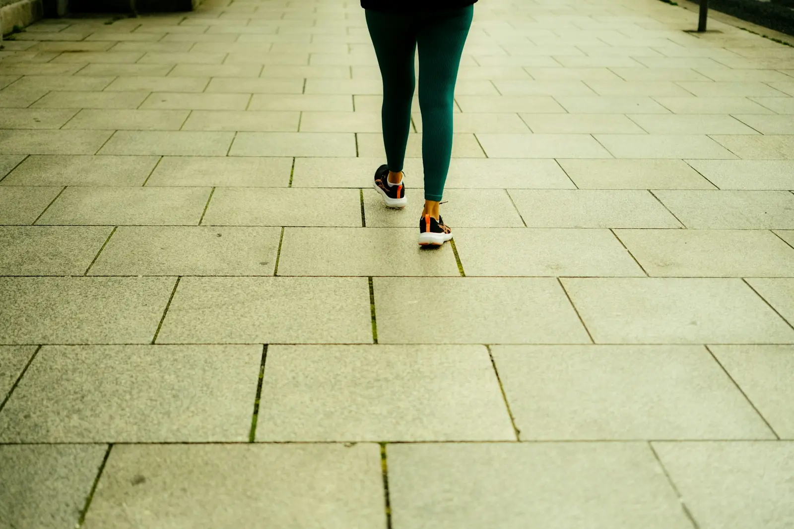 A person's legs in dark green trousers and trainers walking away from the camera along a pavement of large stone slabs