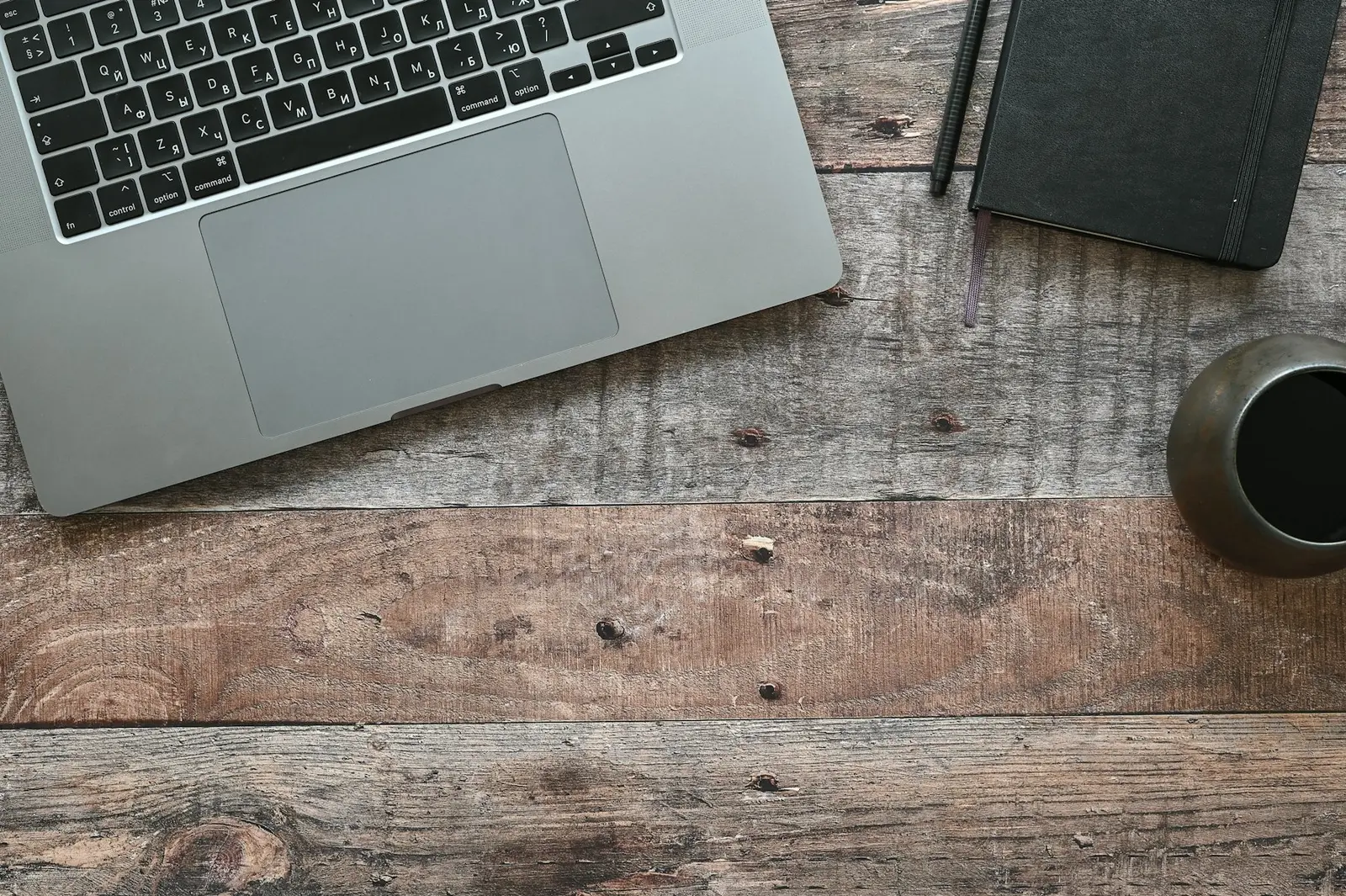 A silver laptop, a black notebook, a pen and a dark cup on a rough wooden desk, seen from above