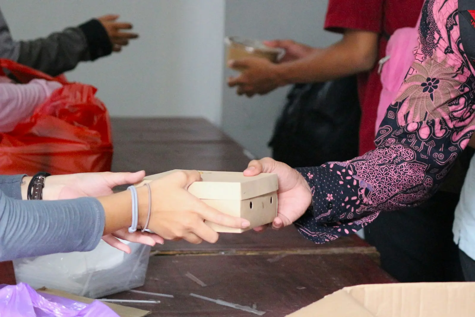 Several hands at a wooden table, one pair passing a small cardboard box to another, with red and purple plastic bags around them