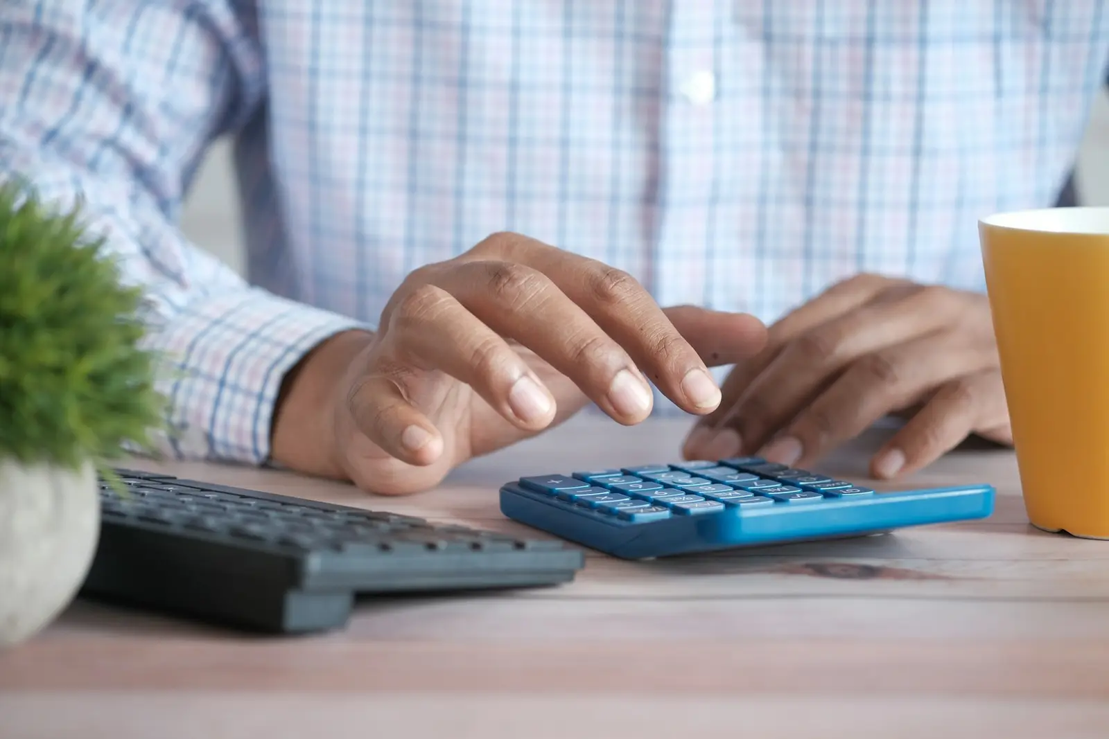 A person in a checked shirt pressing keys on a blue calculator on a desk, next to a keyboard, a small plant and a yellow mug