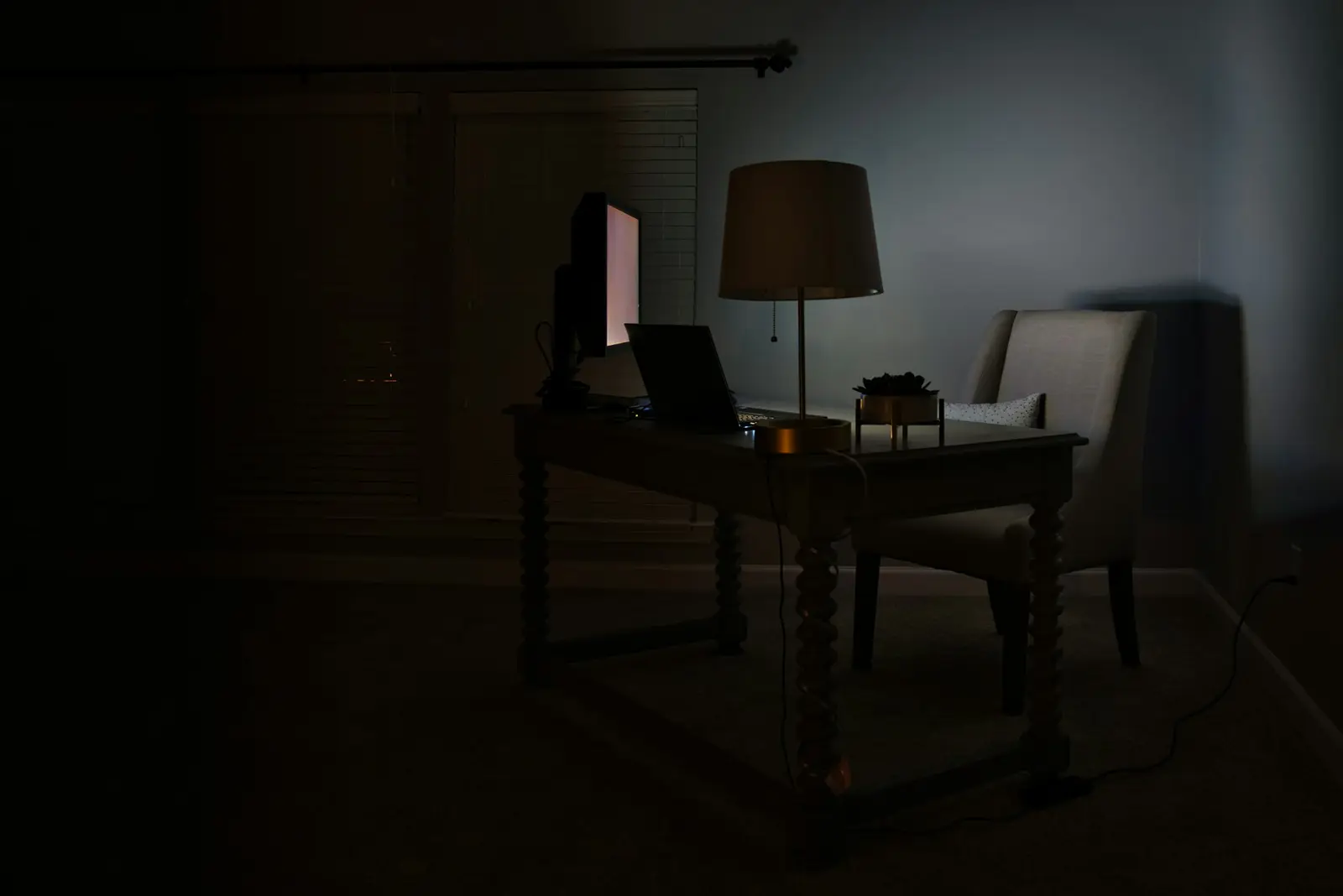 A dim room where a single table lamp glows over a desk and an armchair, with the rest of the room in shadow