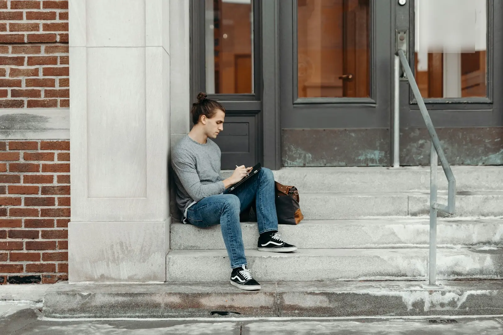 A young man in a grey top and jeans sitting on the stone steps of a building doorway, writing on a tablet on his knees, with a brown bag beside him