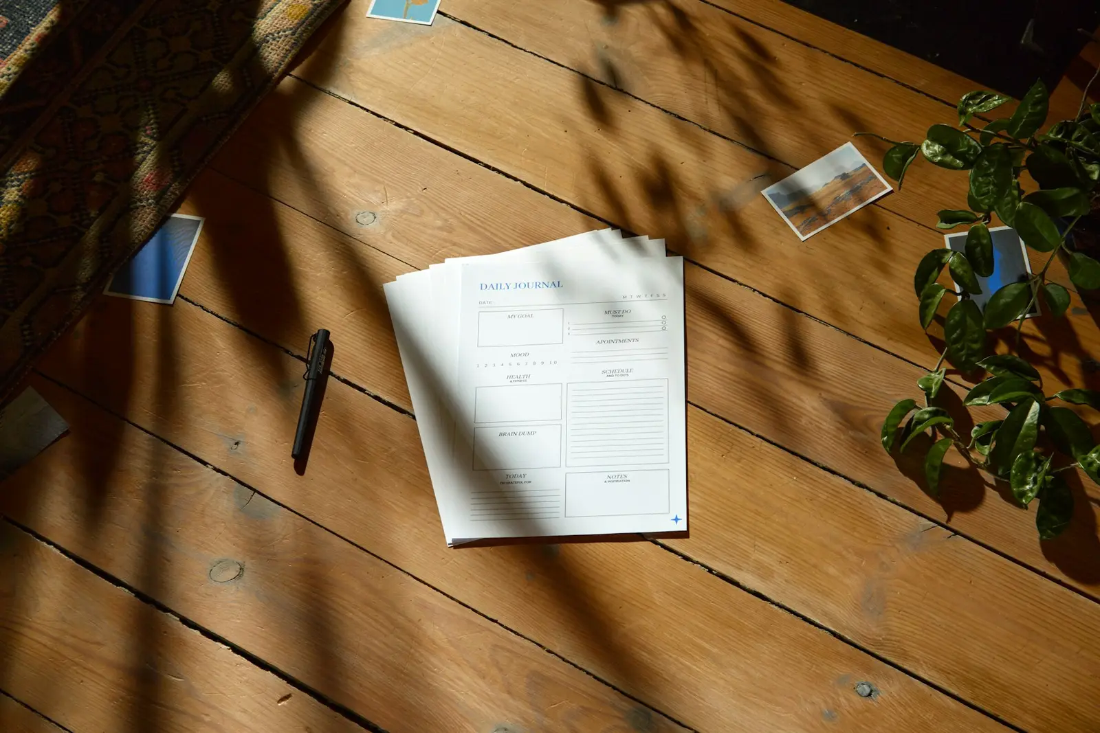 A stack of printed planner pages lying on wooden floorboards in patterned shadow, with a pen and a plant beside it