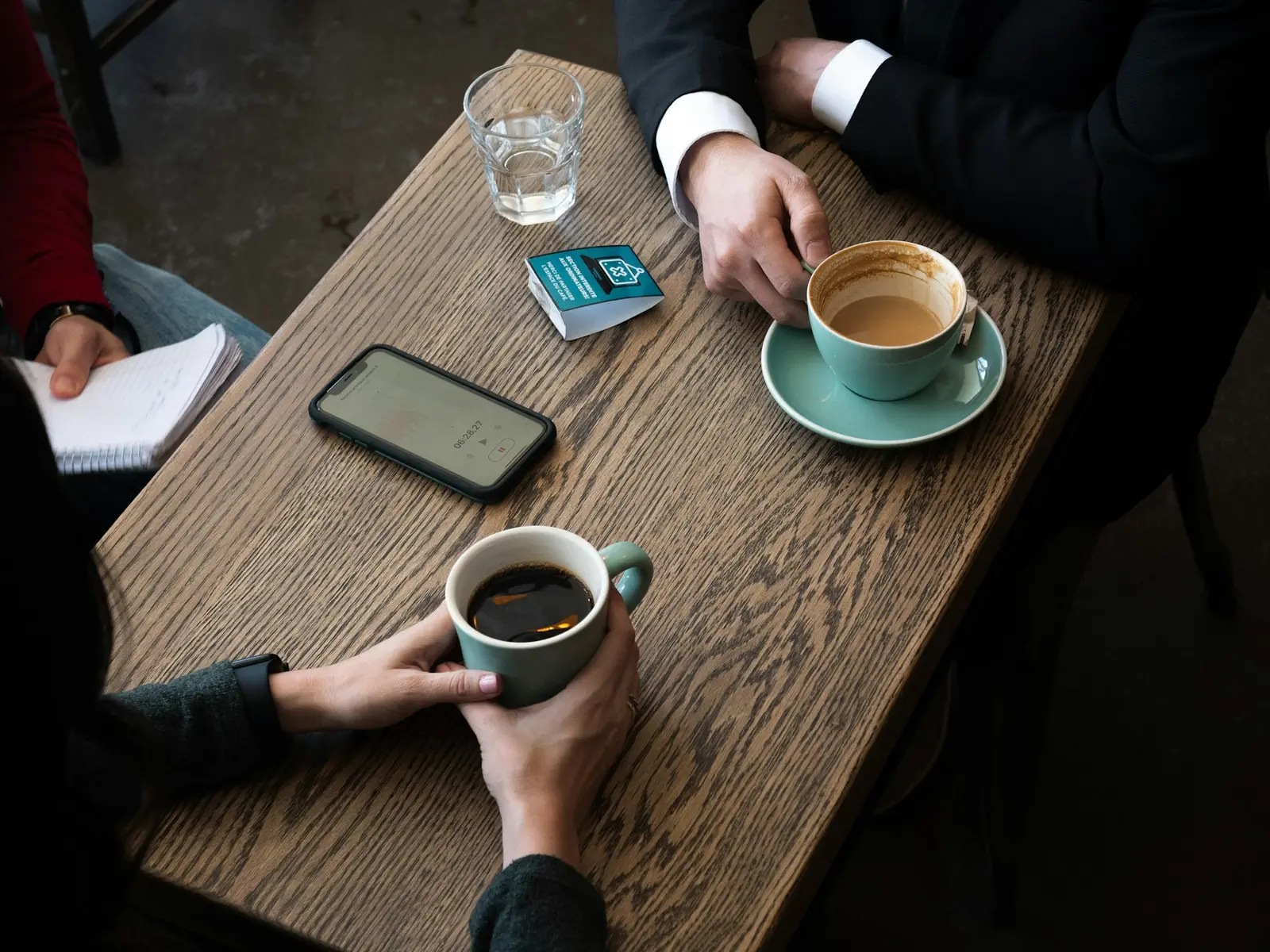 A wooden cafe table seen from above with two cups of coffee, a water glass, a phone lying face up and a card, with a notebook held in one person's hand and other hands around the table