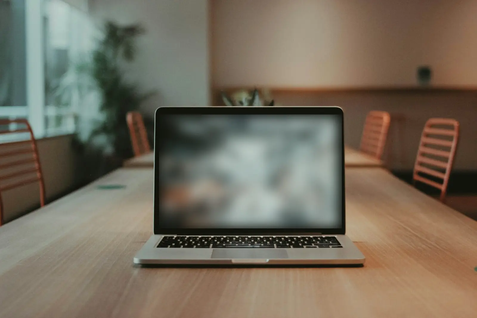 An open laptop on a long wooden table in an empty room, with orange chairs along both sides; the screen has been blurred so nothing on it is readable