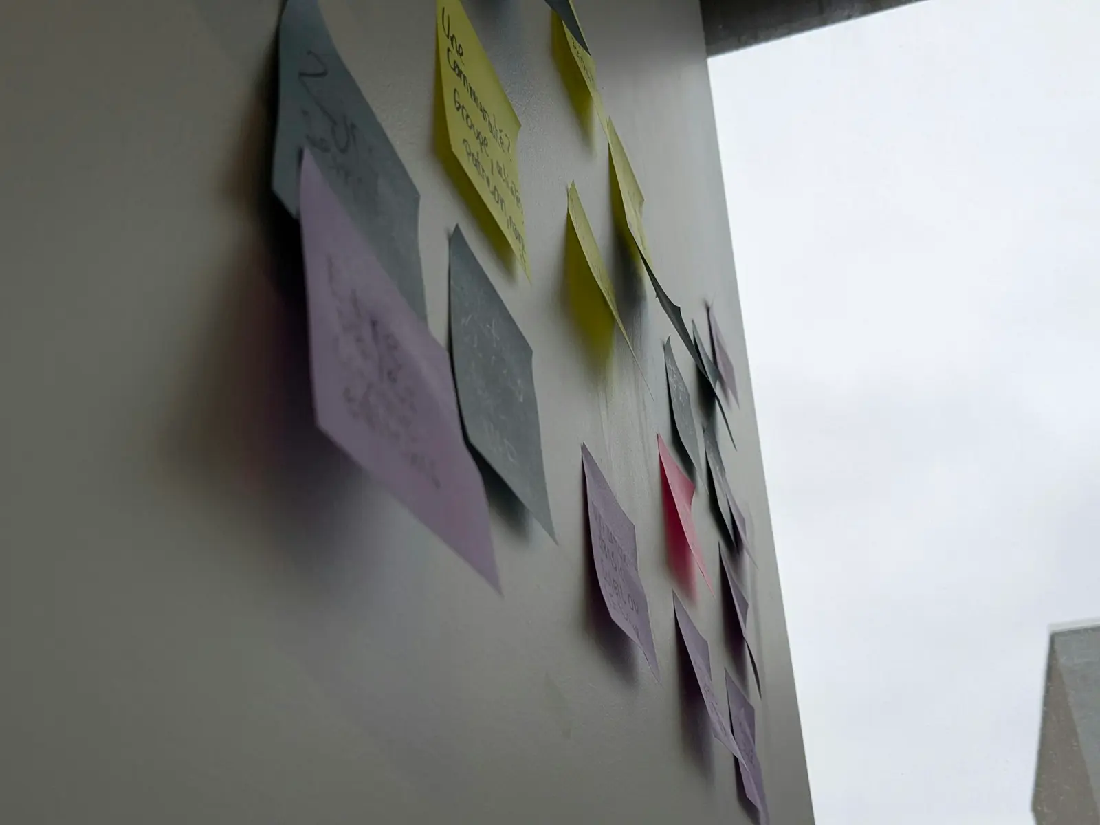 Coloured sticky notes on a grey wall, photographed at a steep angle, the handwriting mostly blurred