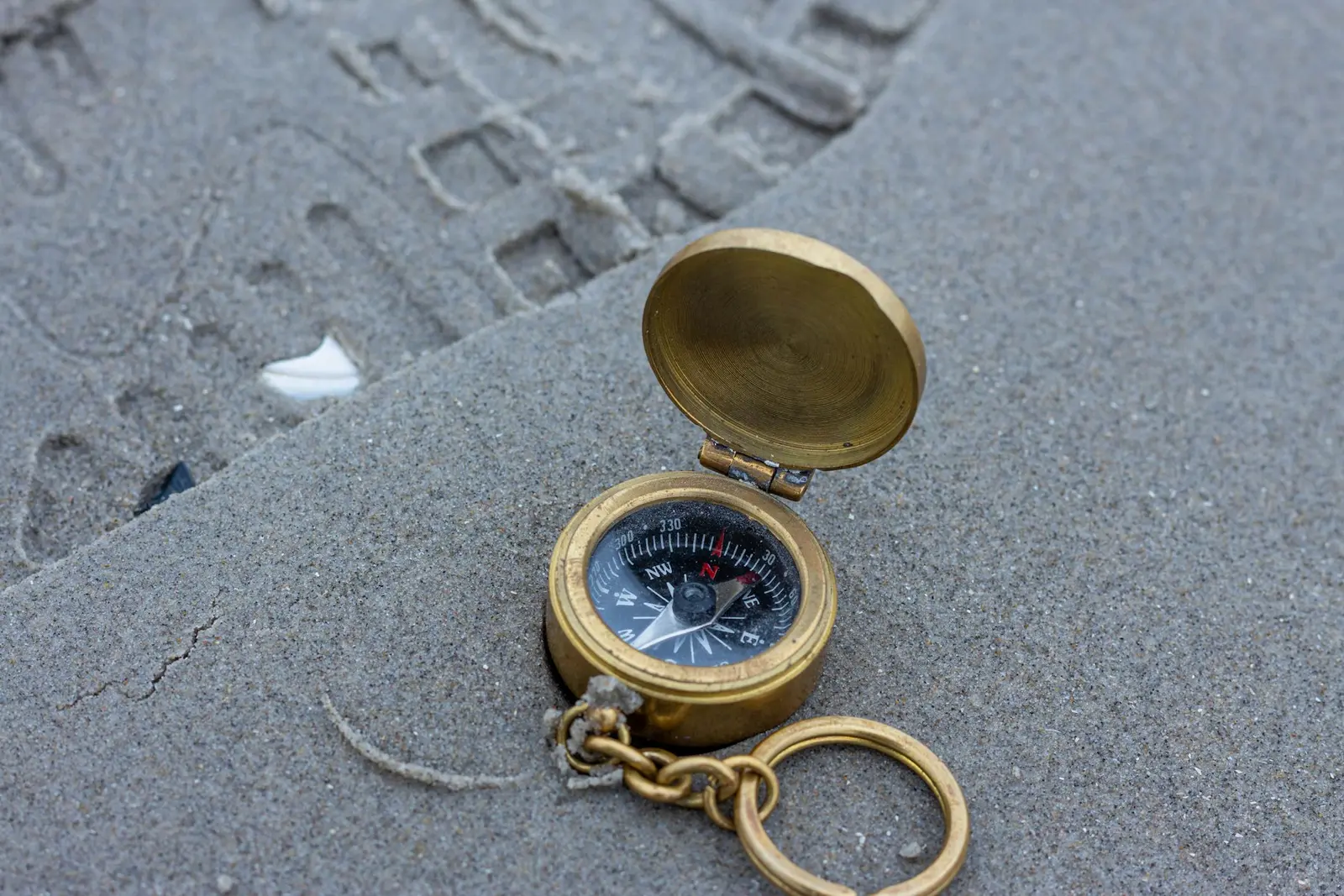 A small brass pocket compass with its lid open, lying on grey sand with a key ring attached
