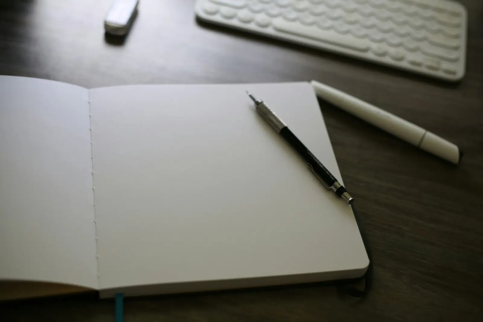 An open blank notebook with a mechanical pencil on it, next to a white pen and a white keyboard on a dark wooden desk