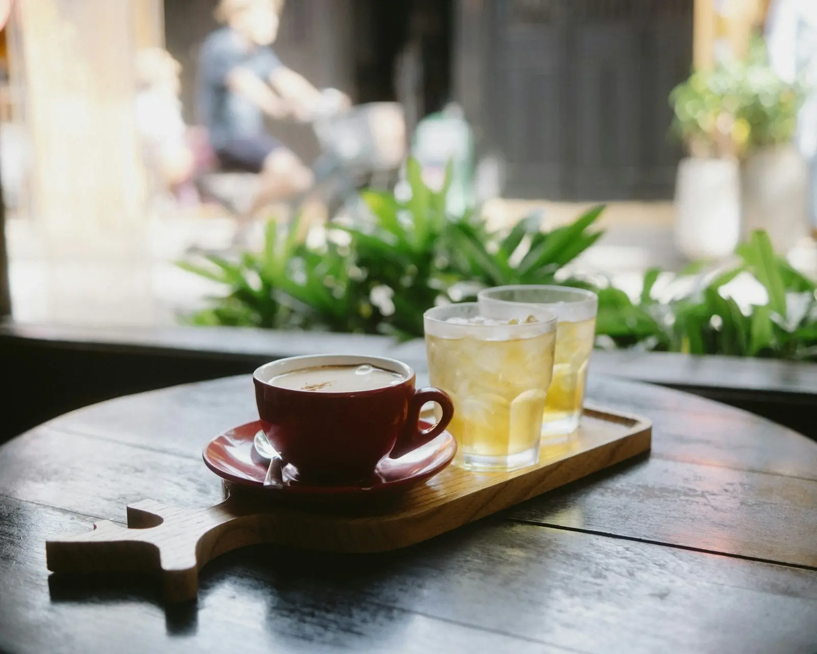 A cup of coffee on a saucer and glasses of iced drinks on a wooden serving board, on a round cafe table with plants behind