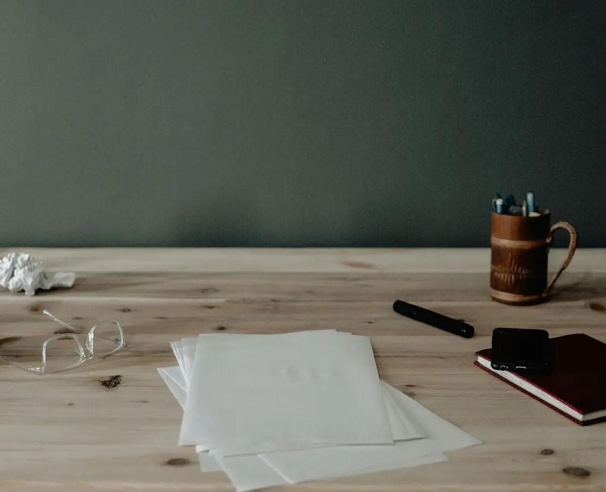 A few loose sheets of blank white paper on a light wooden table, with glasses, a black marker, a pen pot and a red notebook around them