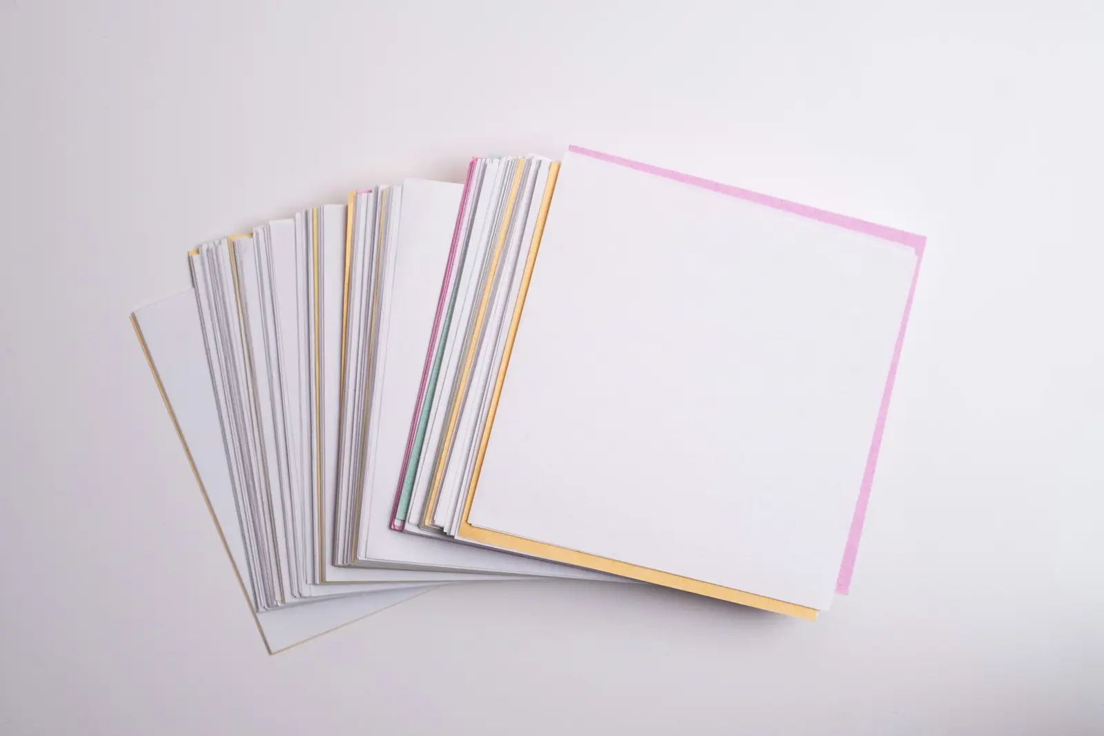 A loose stack of square paper sheets with pink, yellow and blue edges on a plain white surface
