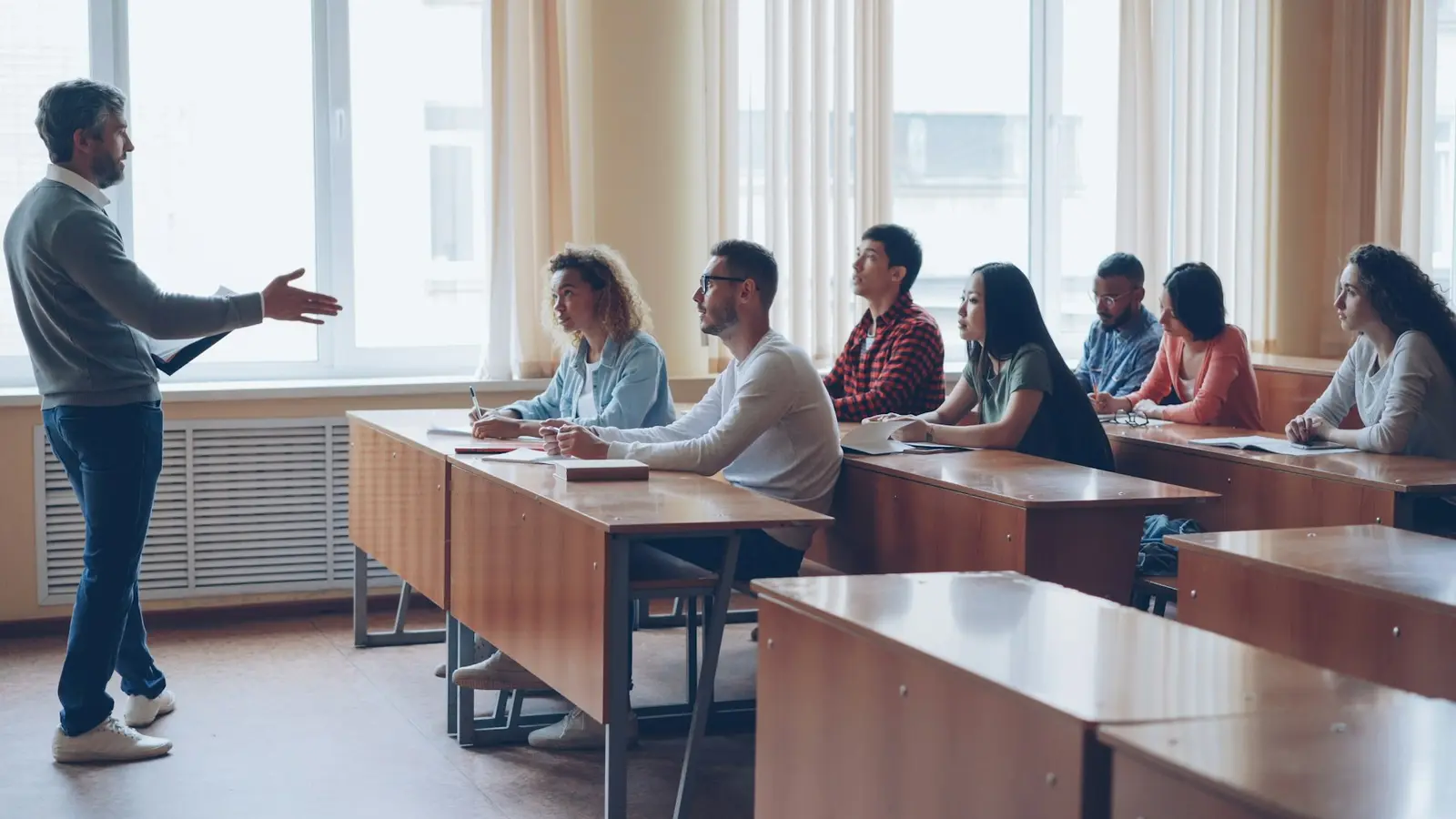 A teacher standing and gesturing to a row of adult students sitting at wooden desks in a bright classroom