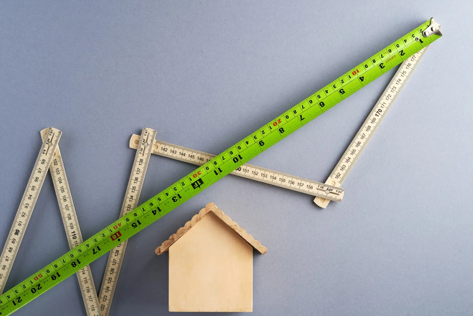 A green tape measure running diagonally across a folding ruler that zigzags over a grey-blue surface, beside a small house-shaped block