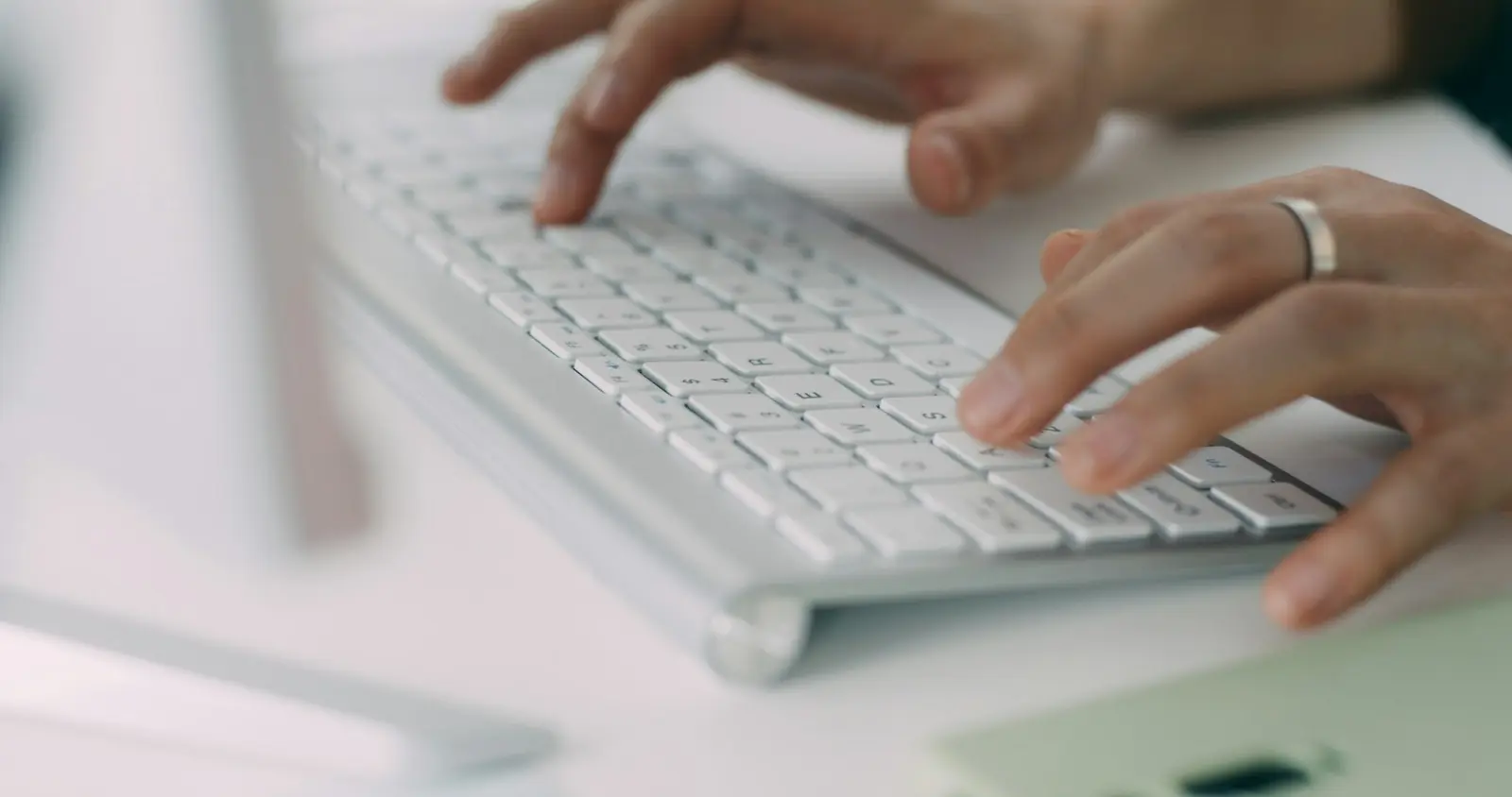 Two hands typing on a white keyboard with the edge of a monitor out of focus beside it, nothing readable on screen