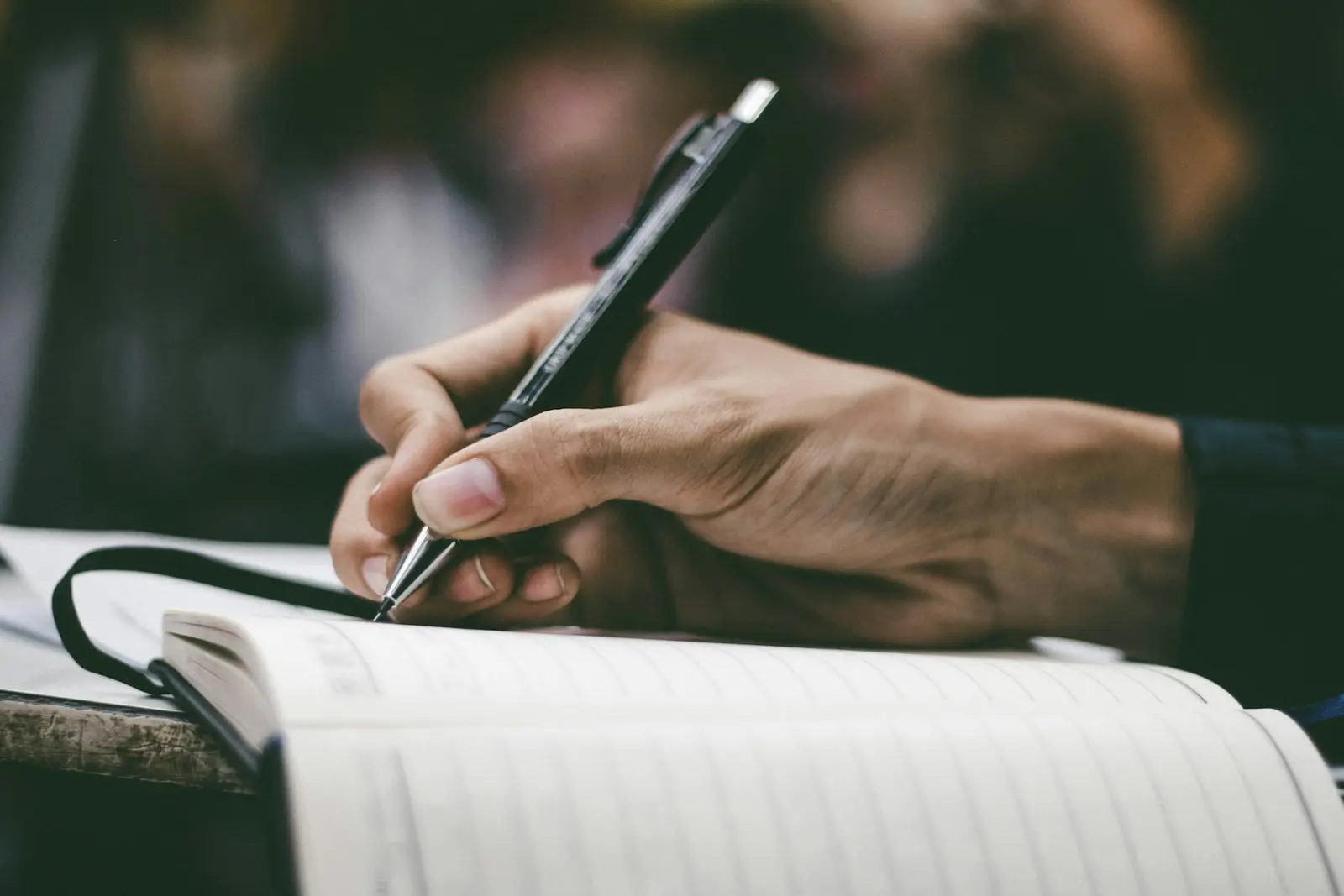 A hand holding a clip-top mechanical pencil over an open ruled journal, writing a line, with the desk edge and a blurred background behind it