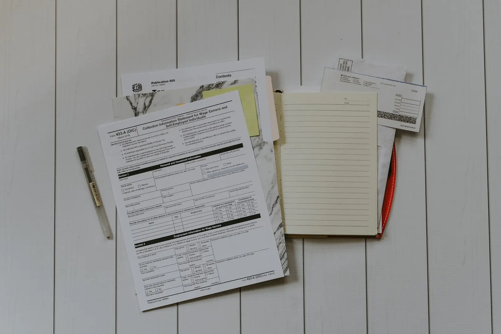 A flat lay of a blank printed government form with rows of Yes and No tick boxes, beside a notebook, a pen and a few envelopes on white wooden boards
