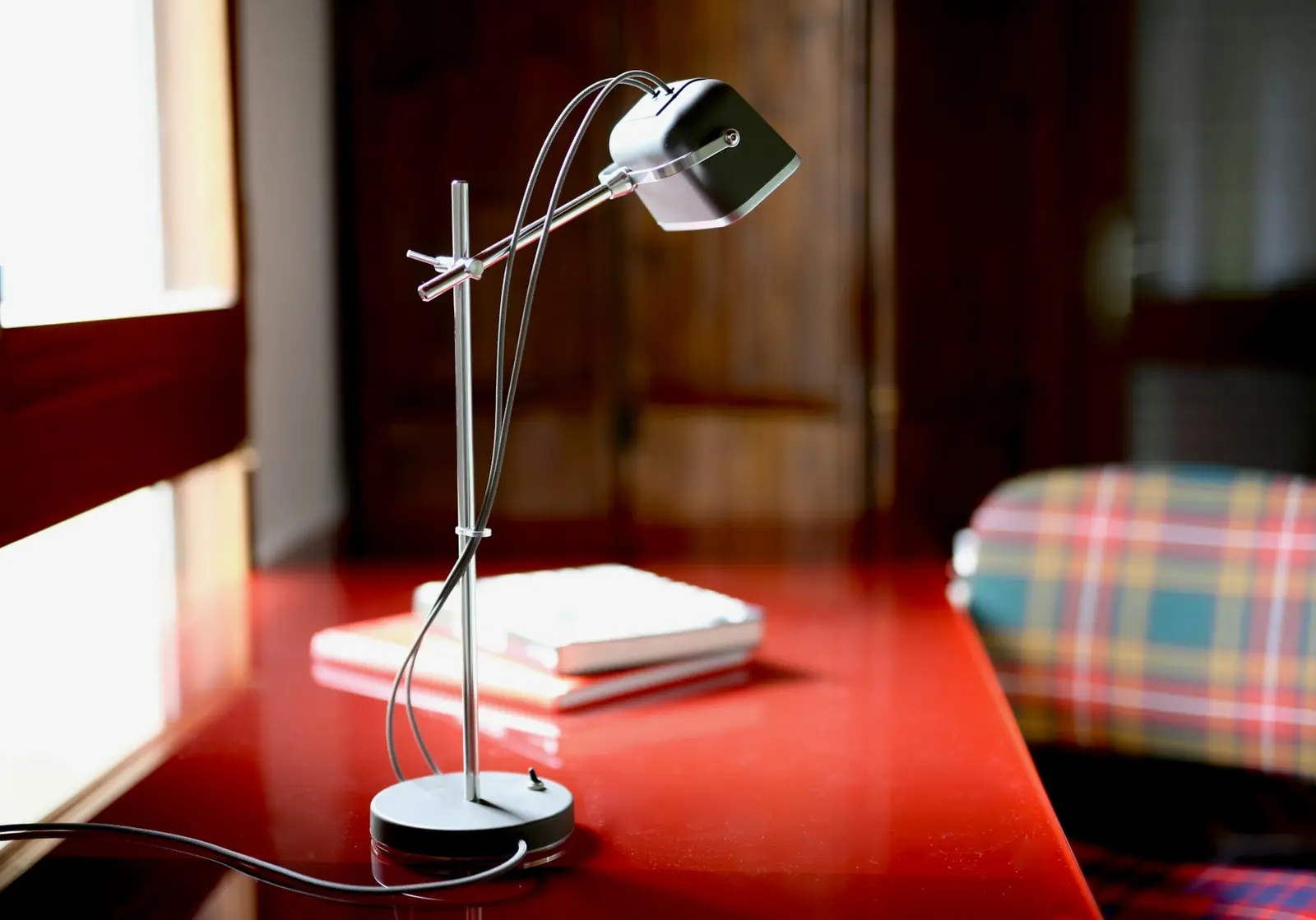 A silver table lamp with a grey head on a red desk beside a window, with a short stack of notebooks next to it and a plaid upholstered chair at the right edge of the frame