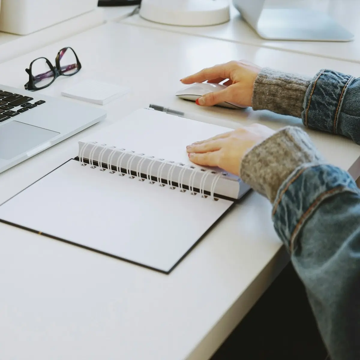 An open spiral notebook and a pair of glasses on a white desk beside the edge of a laptop keyboard, with one of a person's hands on the notebook and the other on a mouse