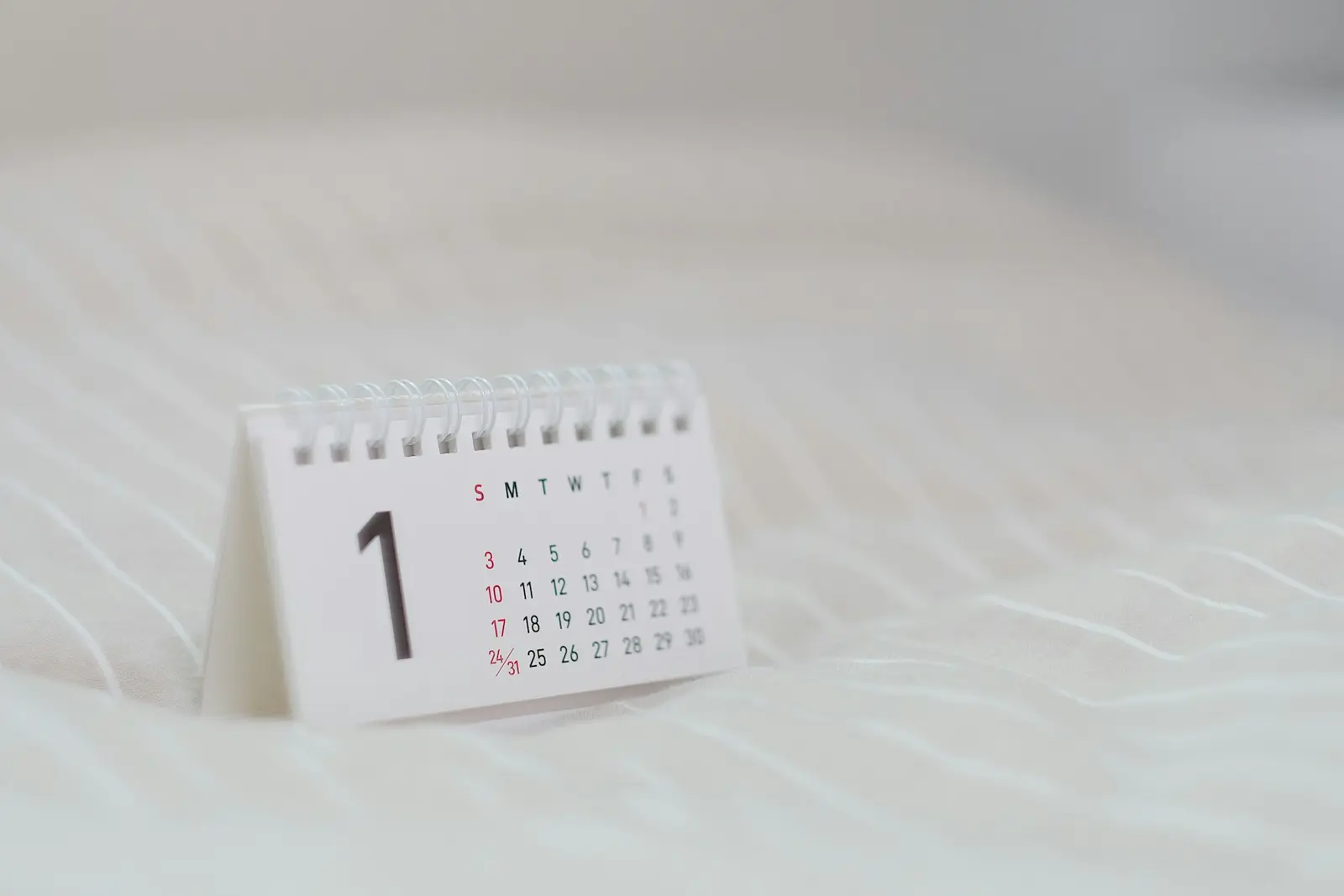 A small white desk calendar with a large number 1 and a grid of dates, standing on a white quilted cloth