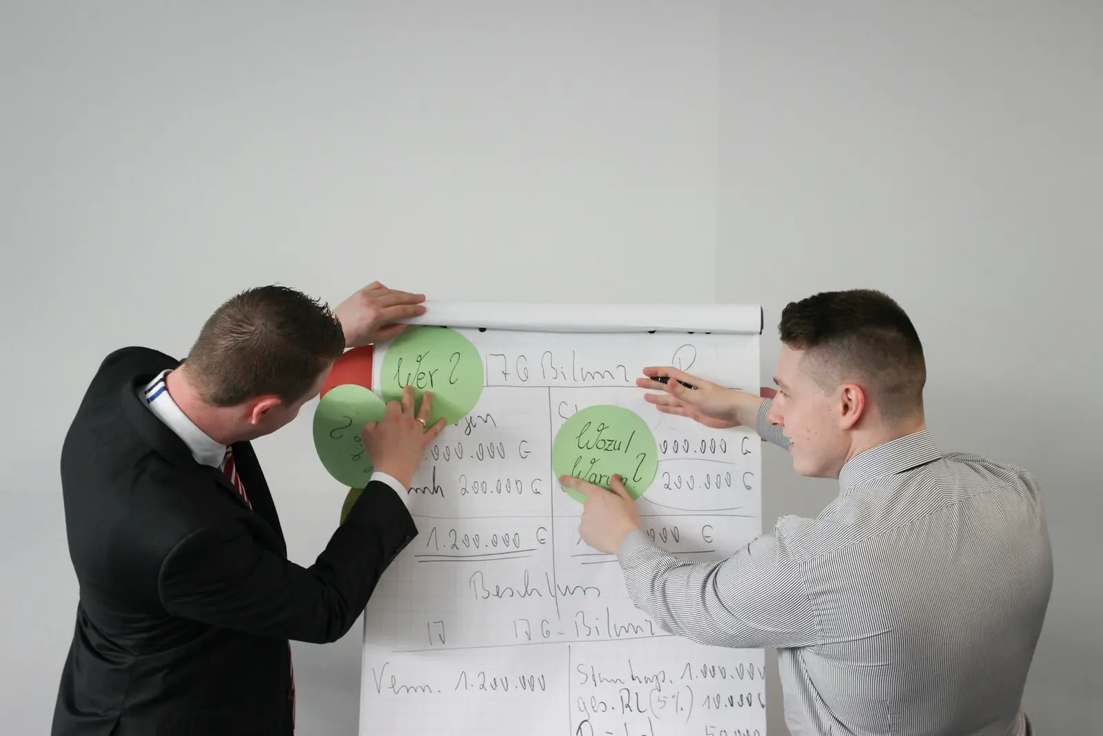 Two men in shirts sticking green paper circles onto a flip chart covered in handwritten figures