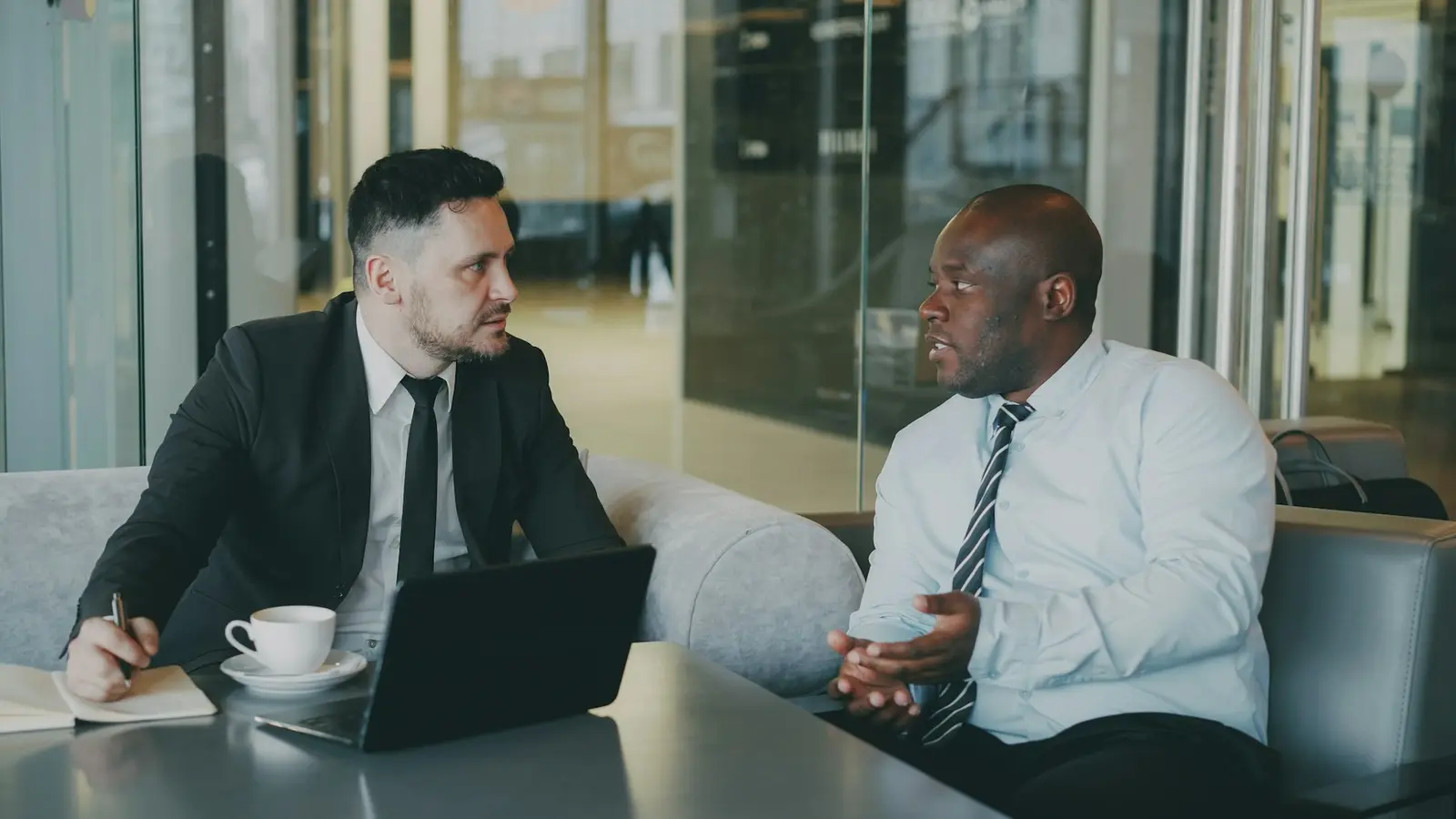 Two men talking at a table in a glass-walled lounge: one in a dark suit with a laptop, a coffee cup and a small notebook in front of him, the other in a light blue shirt and striped tie gesturing as he speaks