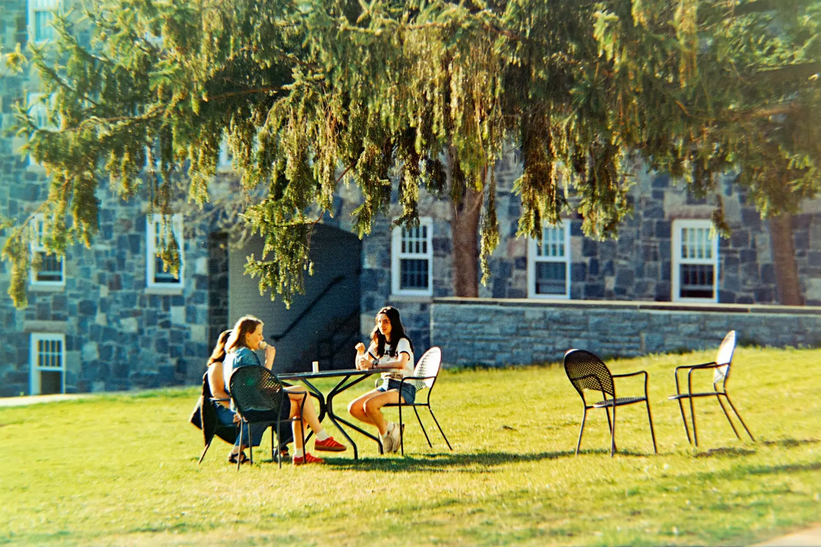 Three people sitting at a small metal table on a lawn under a large evergreen tree, with a stone building behind them