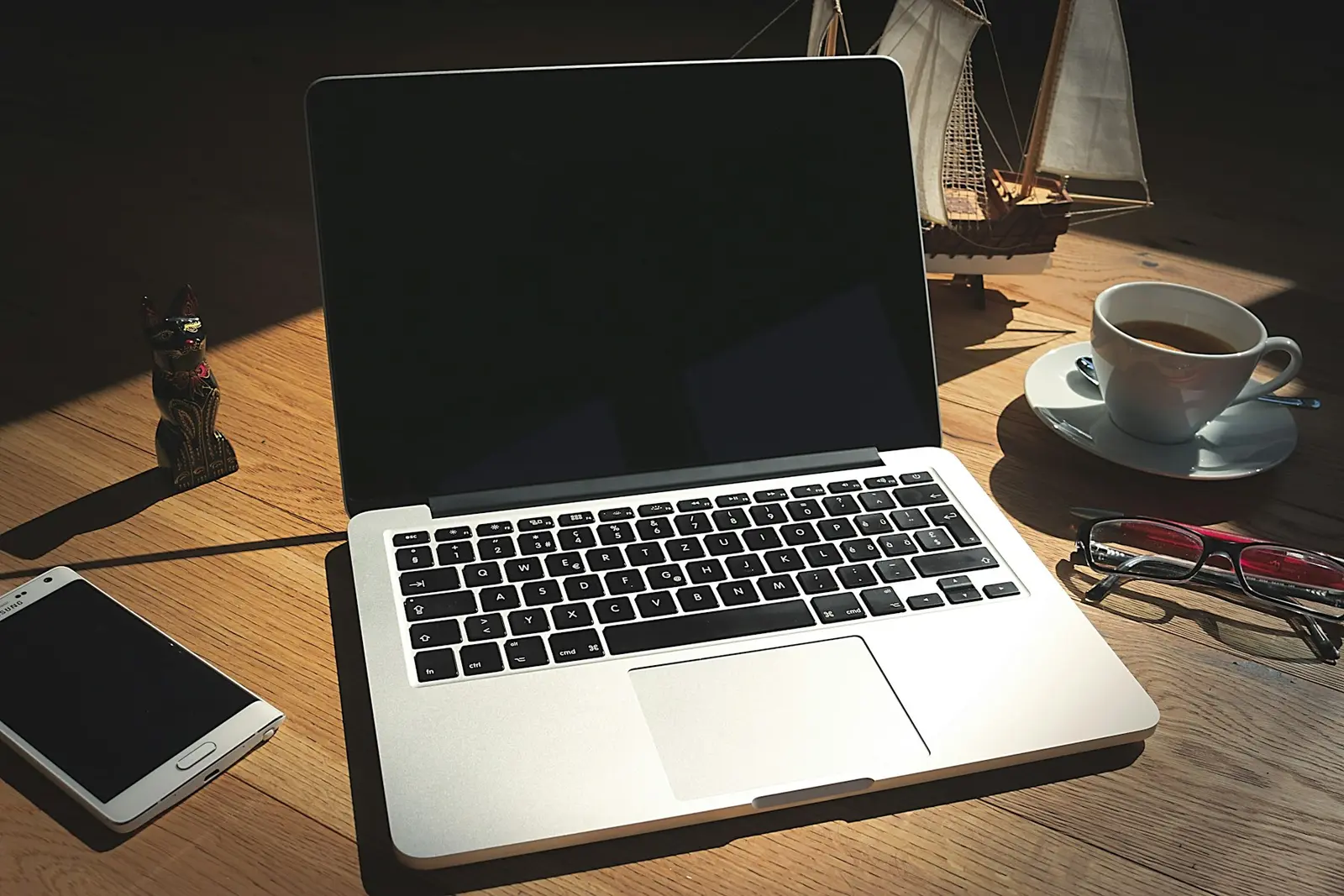 An open laptop with a dark screen on a wooden desk, beside a cup of coffee, a smartphone and a pair of glasses