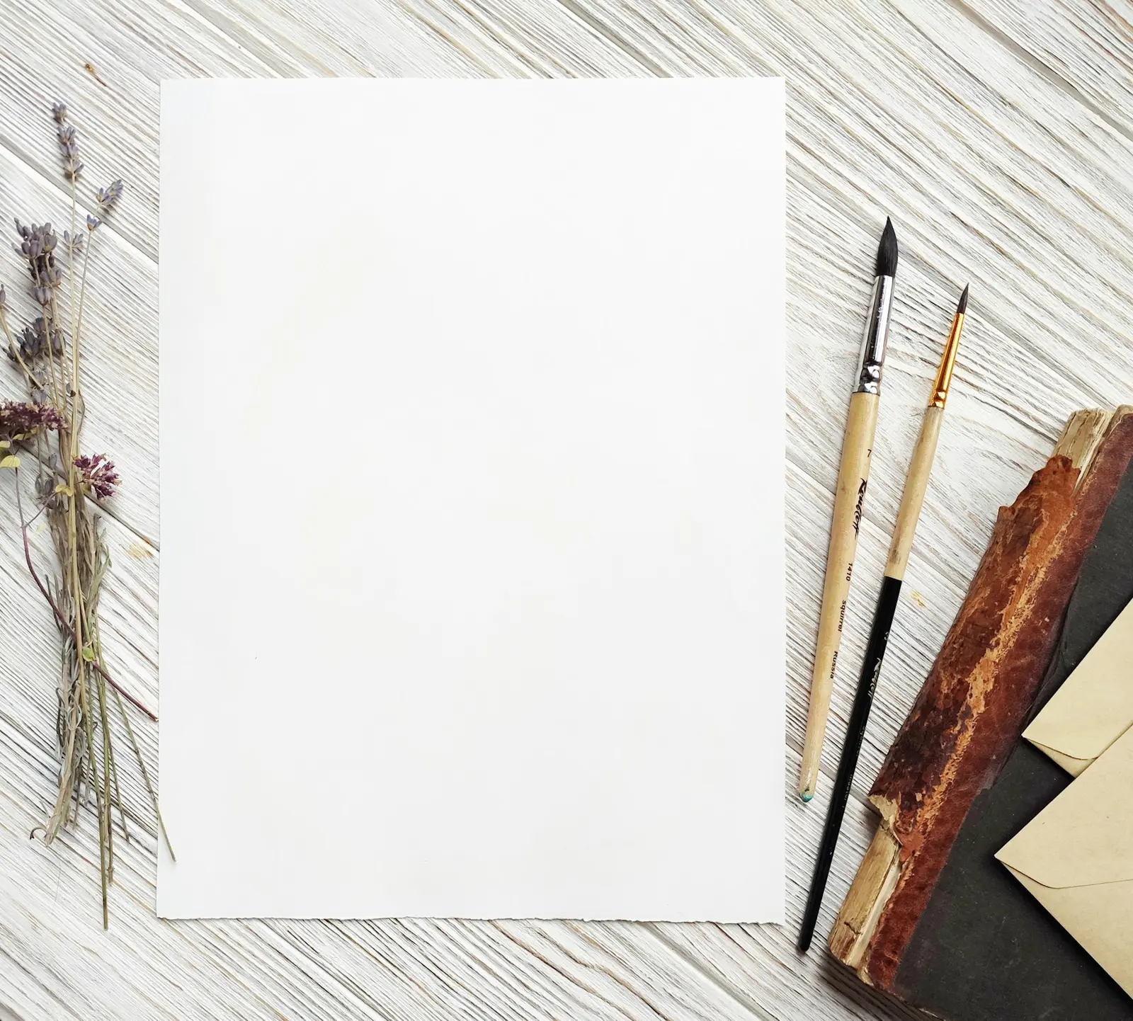 A single blank sheet of white paper on a pale wooden surface, with two paintbrushes, dried lavender and a brown envelope at its edges