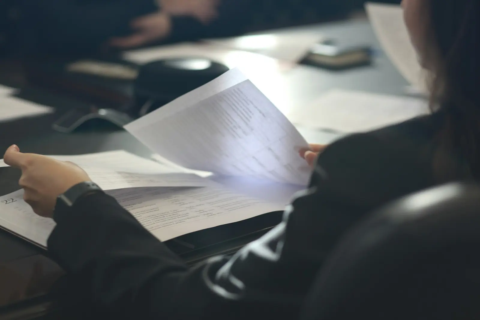 A person in a dark jacket holding up a printed page of small text in a dim room, with more papers spread on the table in front of them