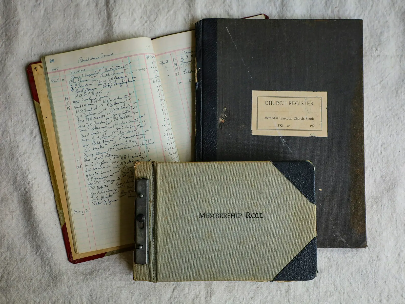 Three old bound record books on a linen cloth: an open handwritten ledger, a dark Church Register and a grey Membership Roll