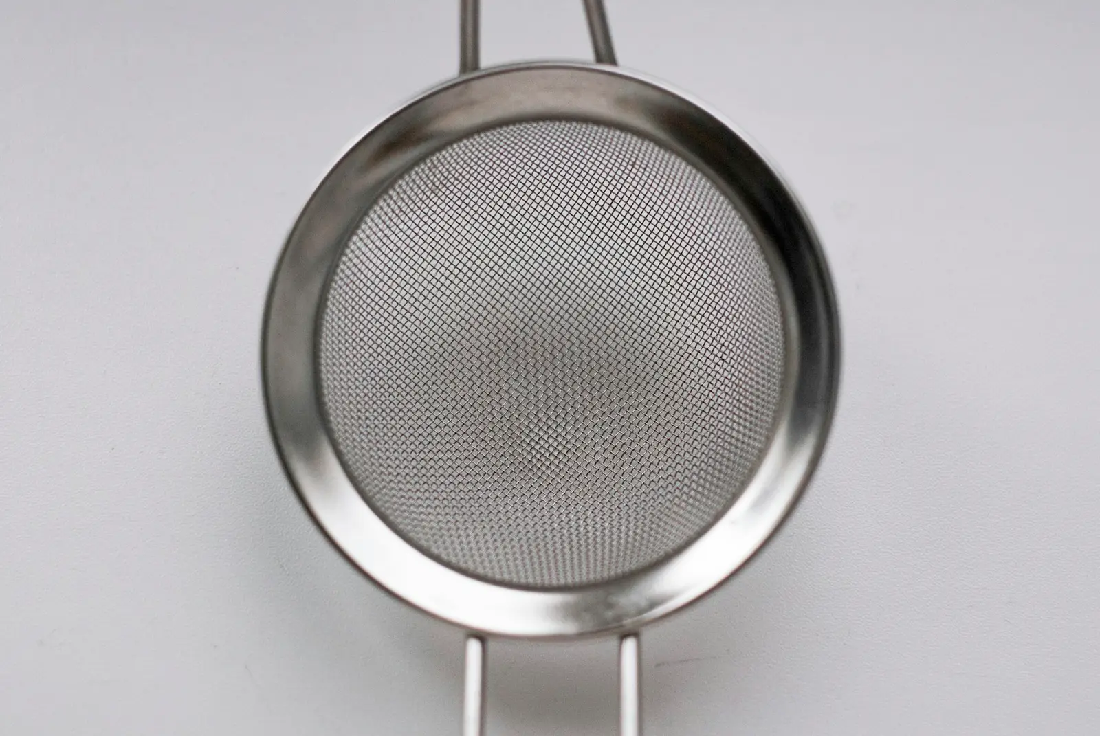 A round stainless steel mesh strainer seen from the front against a plain pale grey background, its handle running off the top of the frame