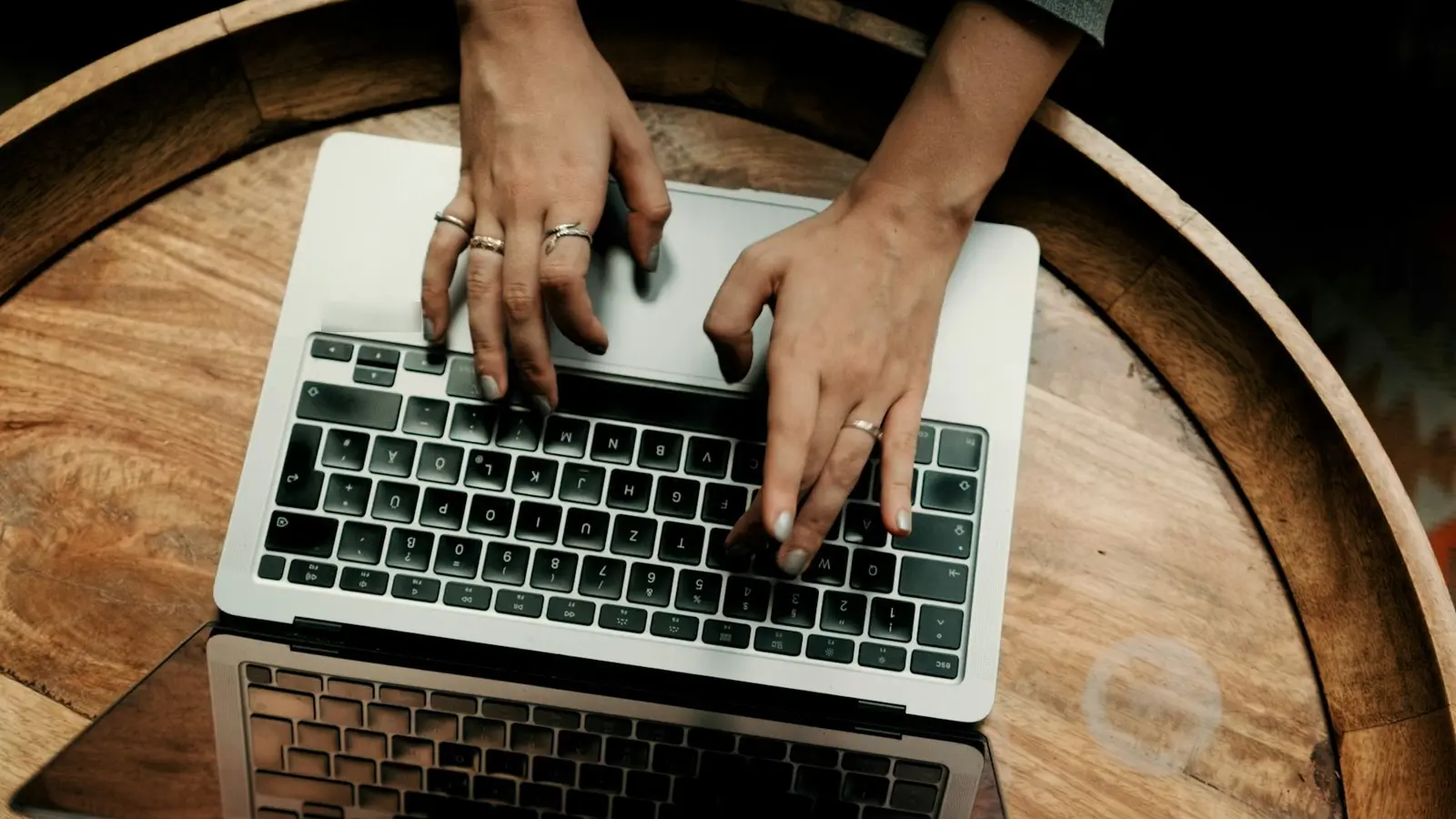 Two hands with rings typing on a silver laptop, seen from above, on a round wooden table