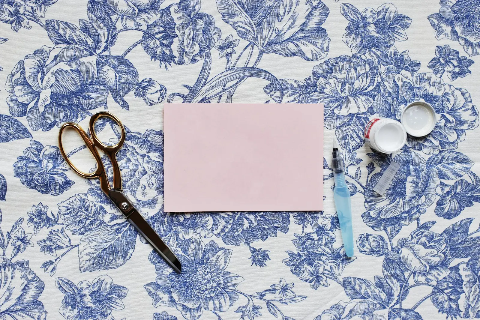A pair of gold-handled scissors, a blank pink card, a water brush, and a small white jar with its lid and a clear cap beside it, laid out on a blue and white floral cloth