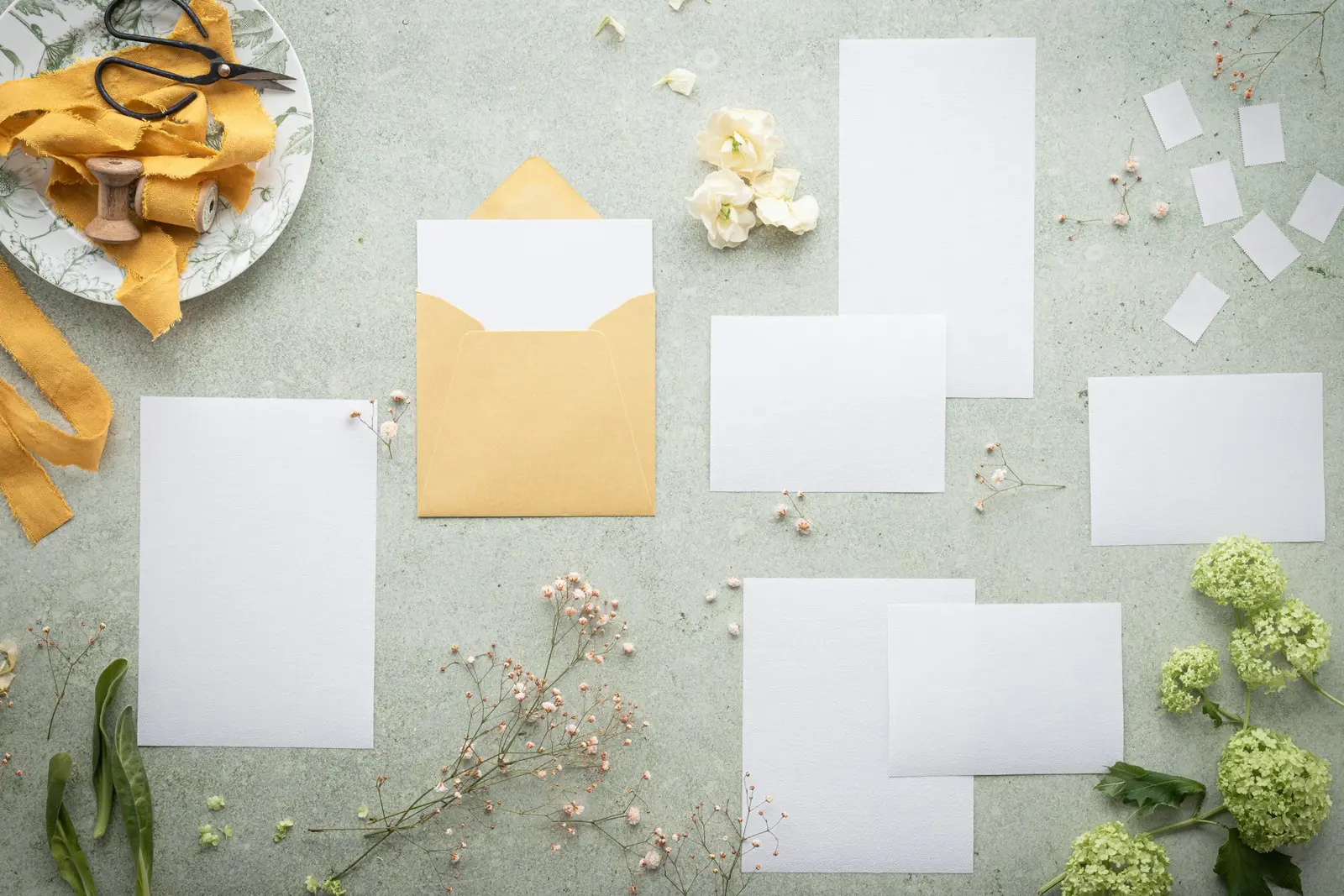 Several blank white cards and an open envelope laid out on a pale green surface, with a plate holding mustard-yellow ribbon on a wooden spool and a pair of scissors at the top left, and small flowers and sprigs around them
