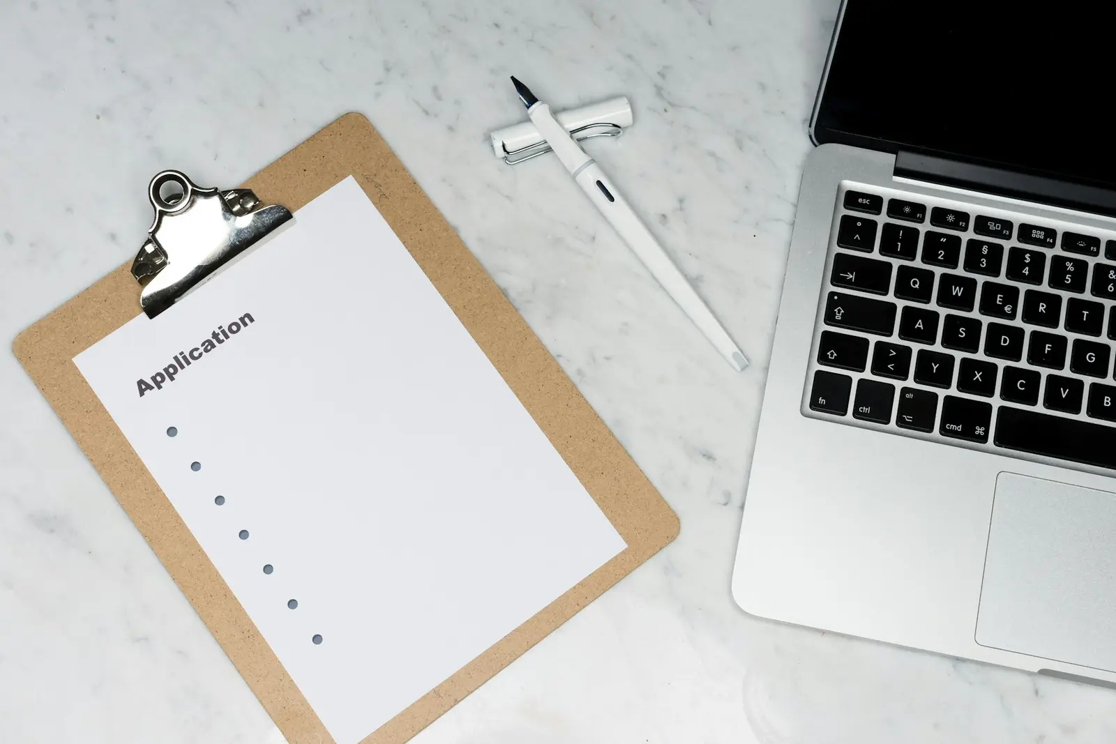 A clipboard holding a sheet titled Application with a column of empty bullet points, next to a pen and an open laptop on a marble desk