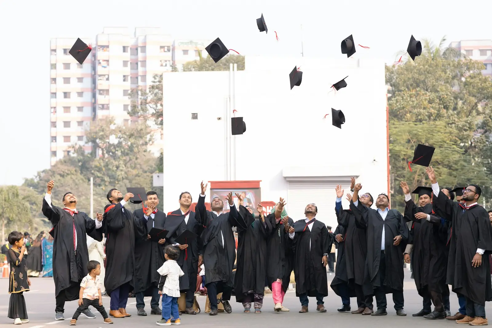 A large group of graduates in black gowns throwing their caps into the air in front of a white building and tall apartment blocks, with a few children standing among them