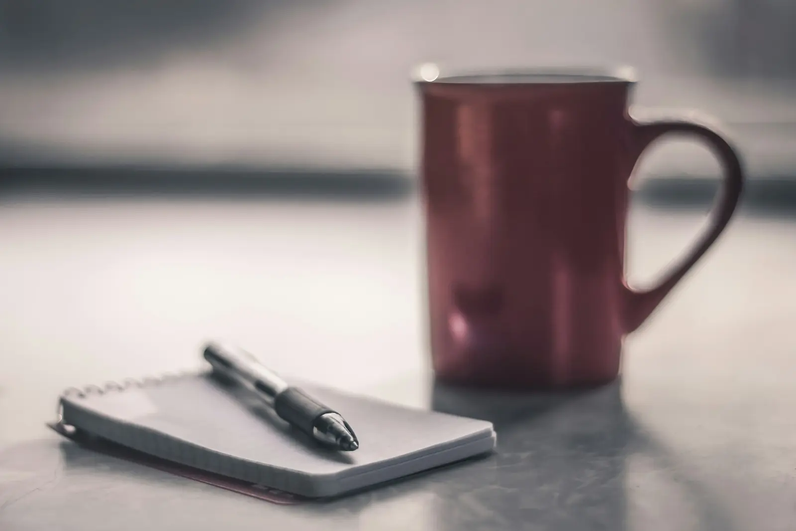 A small notebook with a pen lying on it in front of a dark red mug, on a pale surface