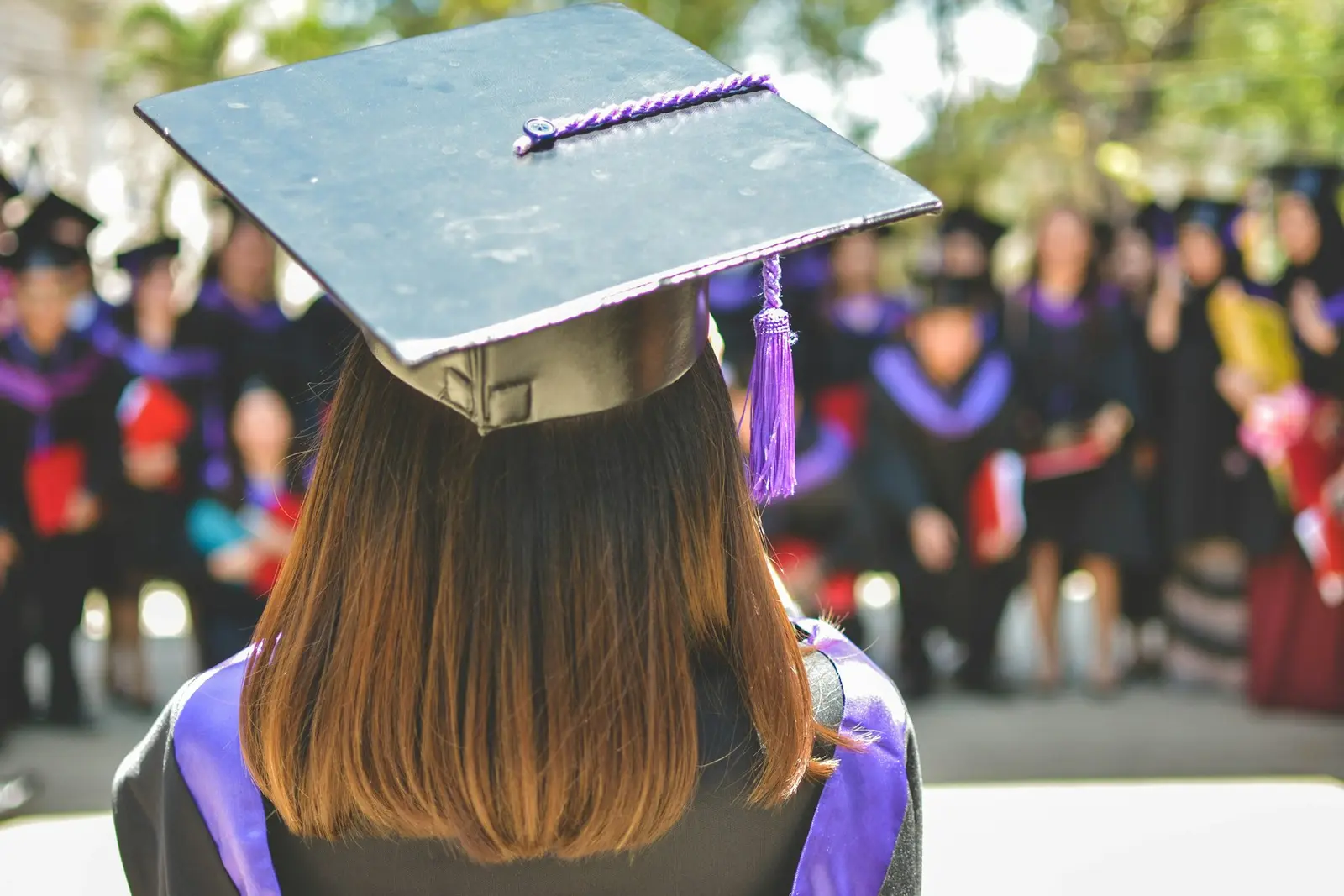 The back of a graduate wearing a square cap with a purple tassel, facing a blurred crowd of other graduates in gowns