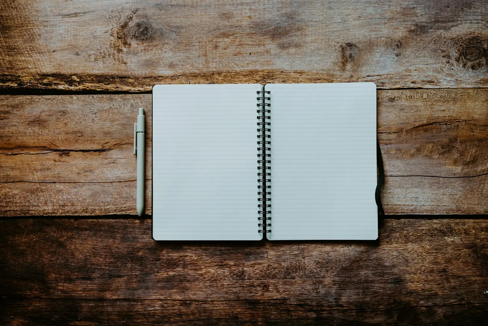 An open spiral-bound notebook with blank lined pages lying on a worn wooden table, with a pale grey pen beside it