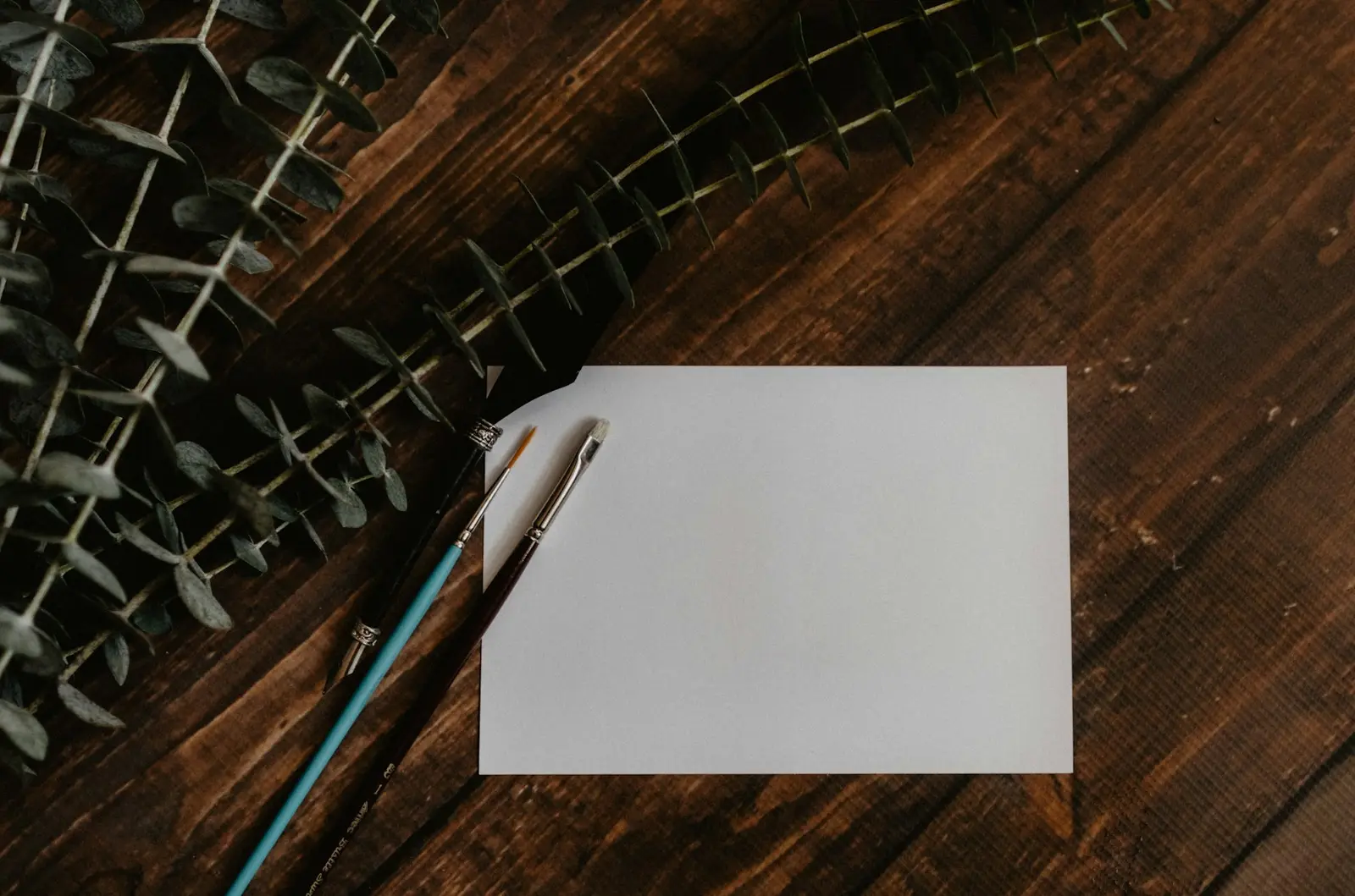 A blank white card and two pens on a dark wooden table, with sprigs of eucalyptus at the edge