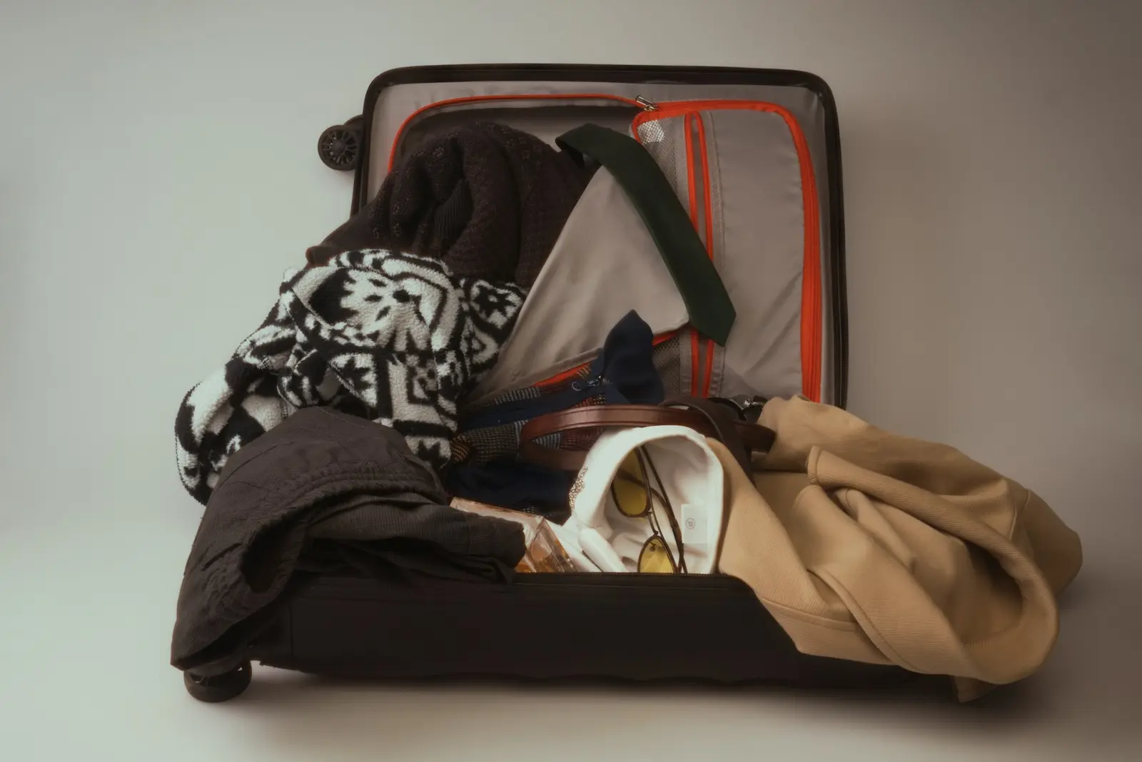 An open suitcase overflowing with clothes, a tie, a belt and a beige coat, against a plain grey background