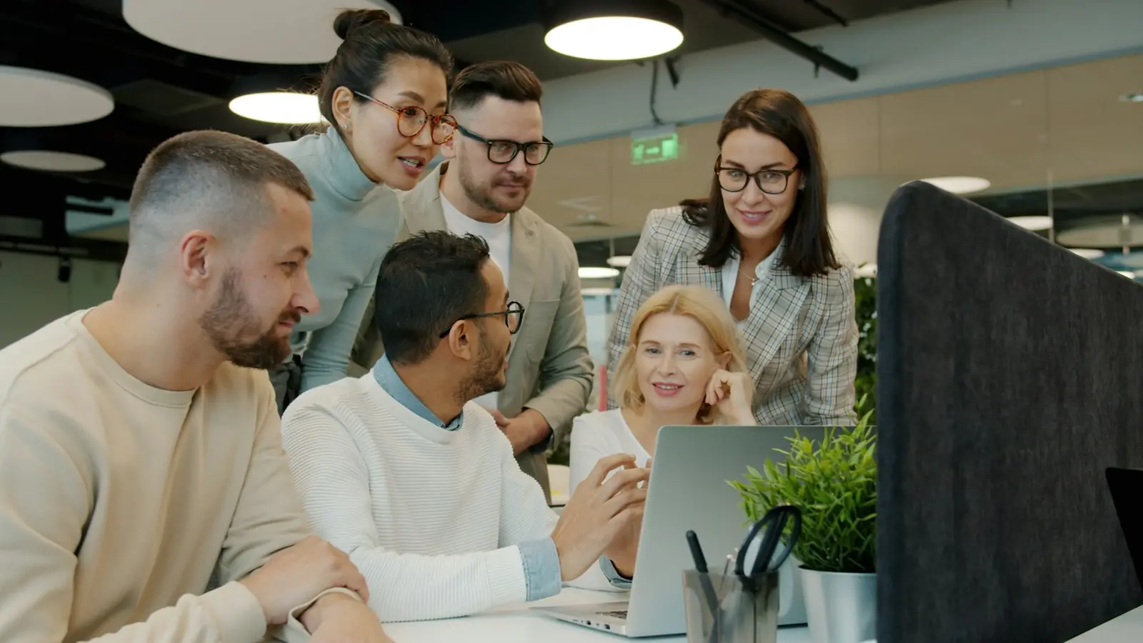 Six colleagues gathered around one laptop in an open-plan office, one man pointing at the screen while the others watch and talk