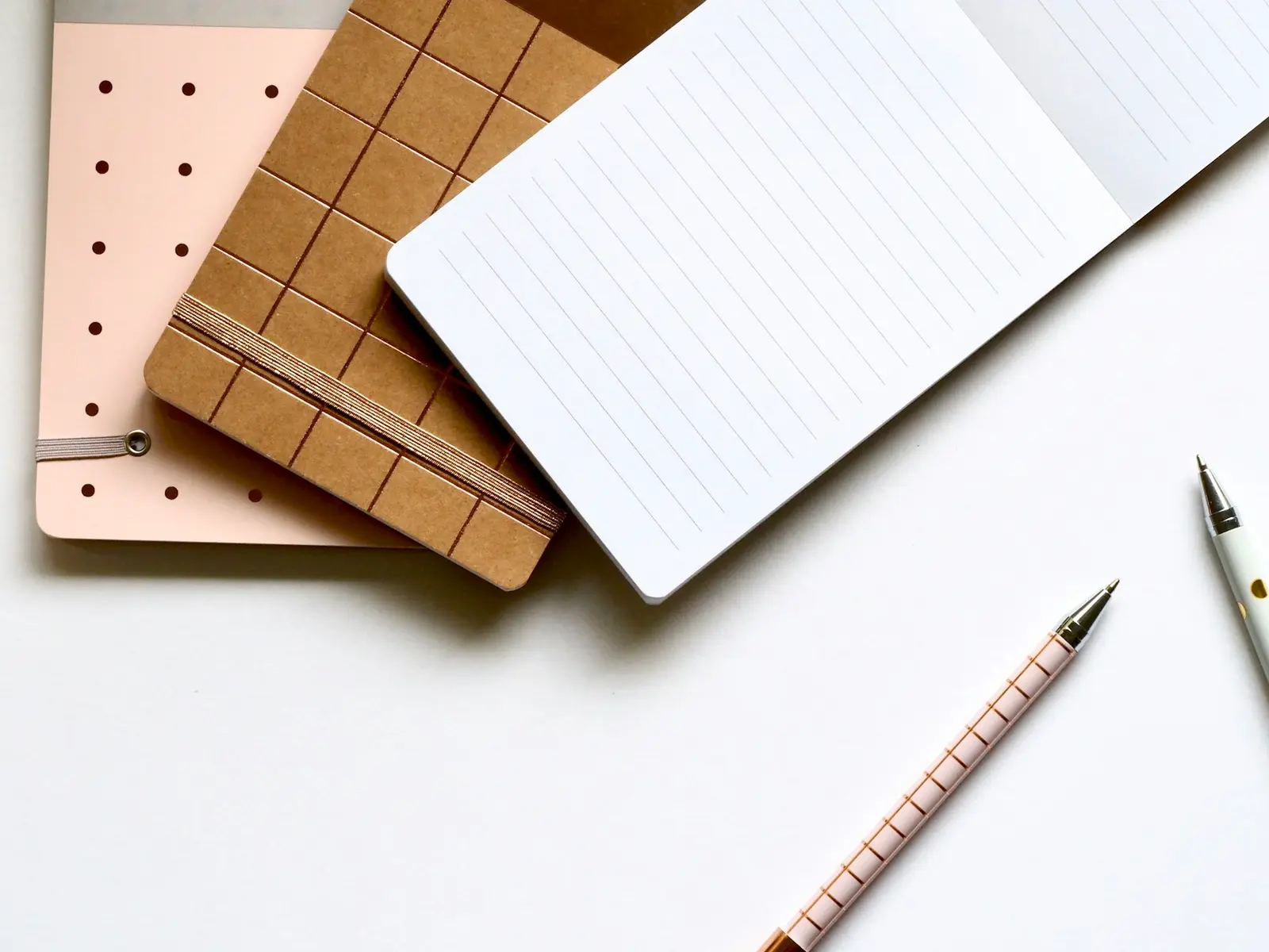 Three notebooks, one pink with dots, one kraft brown with an elastic band and one open with blank lined pages, next to two pens on a white surface