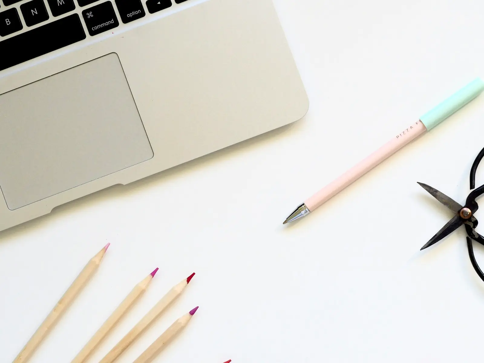 The corner of a silver laptop on a white desk, with a pastel pen, a pair of scissors and three coloured pencils beside it