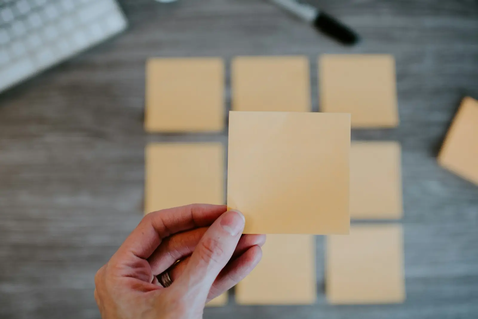 A hand holding up one blank orange sticky note in front of a neat grid of matching blank sticky notes on a grey wooden desk