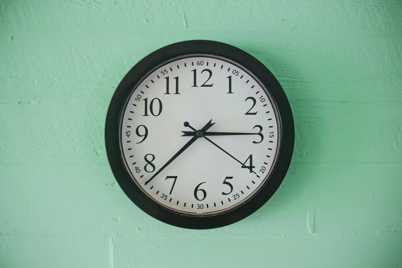 A plain round wall clock with a black frame and a white face, hanging on a mint-green wall