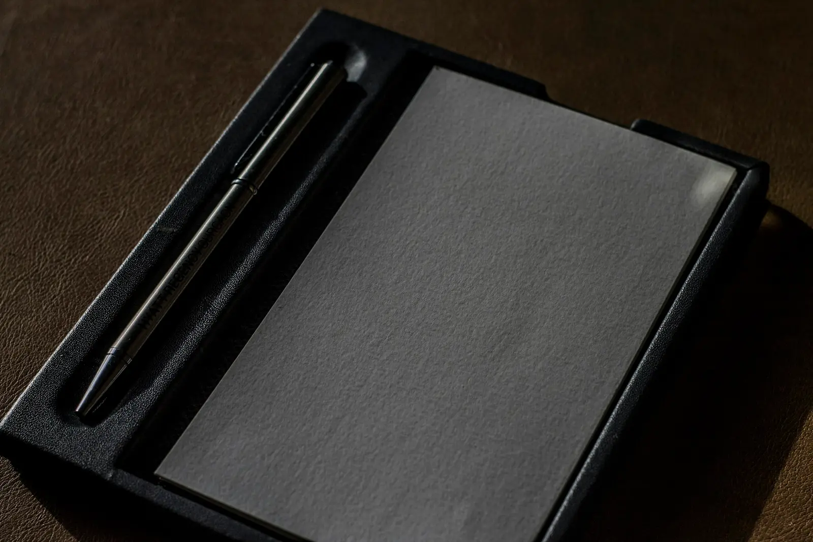 A black pen beside a flat grey rectangle, both set into a black case on a dark surface