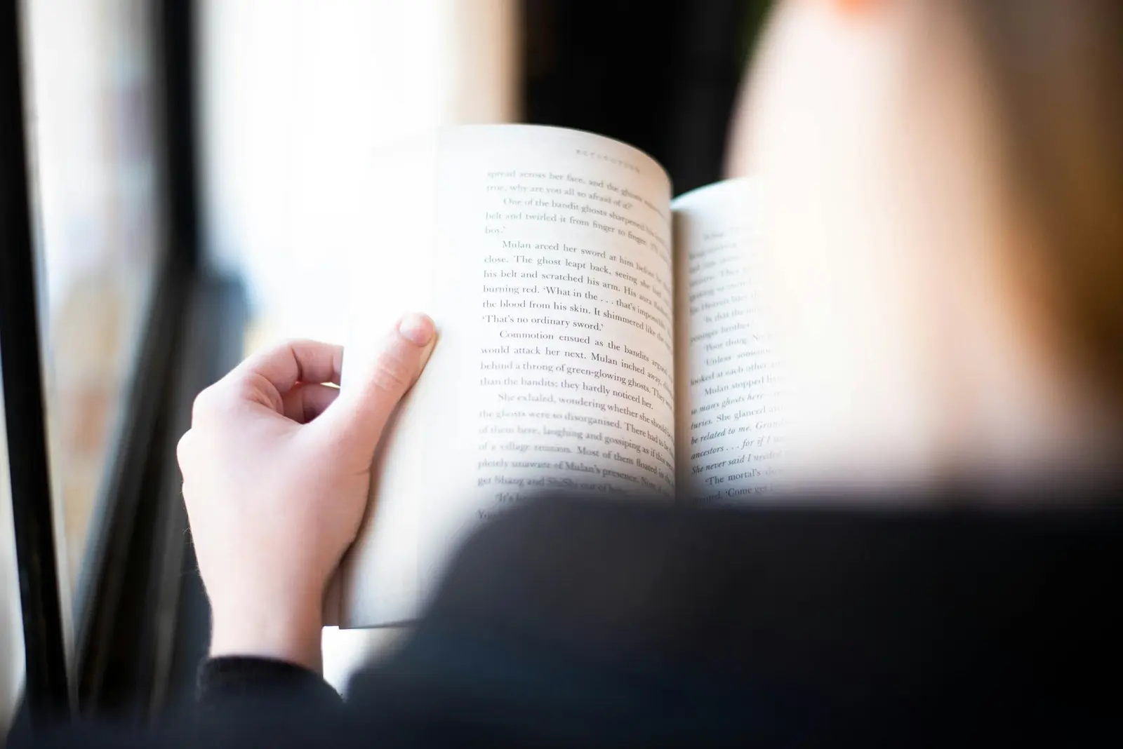 A person seen from behind holding an open printed book up to a bright window, one hand on the edge of the page